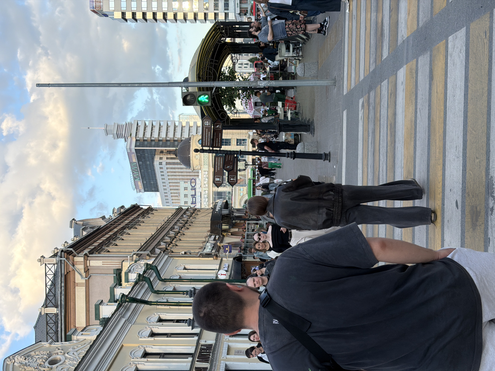 The image presents a detailed view of a bustling pedestrian street in Kazan, Russia. Identification of the location is confirmed by a visible "GRAND KAZAN HOTEL" sign on a building in the mid-ground, and a black lamppost on the left displaying directional signs in both Cyrillic and English, pointing towards "KAZAN UNIVERSITY" and "SOVIET LIFESTYLE MUSEUM." The scene appears to be set in the late afternoon or early evening, evidenced by the warm, diffuse light and a partly cloudy sky.
In the immediate foreground, a male figure is captured from behind, positioned on a yellow and white striped pedestrian crossing. He wears a dark gray short-sleeved t-shirt and light gray shorts, with a black strap from a shoulder bag visible across his back. Slightly to his right and further back, another female figure walks away from the camera, dressed in a dark leather jacket and dark flared pants.
The street is populated with numerous pedestrians walking in various directions. On the right side of the street, under a decorative arched metal structure, a group of people are seated at tables, suggesting an outdoor cafe or gathering area. A green pedestrian traffic light is illuminated on a pole to the right of this structure, indicating permission to cross. A tall, plain grey pole stands vertically in the center of the visual field, behind the foreground figures.
On the left side of the street, a row of ornate, multi-story historical buildings with intricate facades, including decorative elements and green downspouts, lines the thoroughfare. Beyond these historical structures, modern high-rise buildings are visible in the background, including the aforementioned "GRAND KAZAN HOTEL" and a building featuring a prominent dark dome. The pavement is clearly defined with sidewalks and a well-marked pedestrian crossing. The overall environment suggests a vibrant and well-maintained urban center.