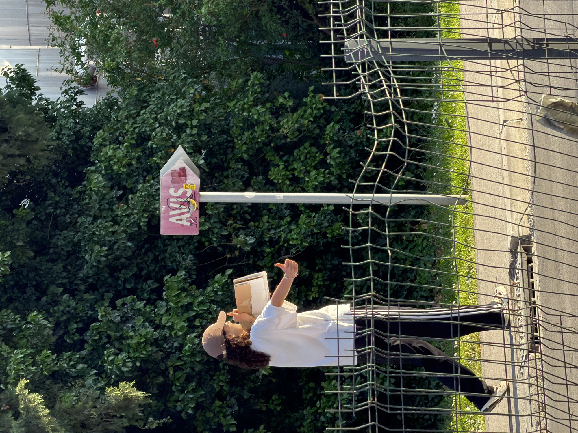 This outdoor image, taken in Budva, Montenegro, depicts a daytime scene featuring a person, a unique sign, and a metal fence set against a backdrop of dense green foliage. A young person, possibly a woman, stands in the foreground, dressed in a white t-shirt, dark pants with white stripes, and a brown baseball cap covering curly dark hair. She holds an open cardboard box or book, intently looking down at it, with her right arm raised and her hand open as if gesturing or reading aloud. A silver bracelet adorns her wrist.
Behind the individual, a profusion of green trees and bushes creates a vibrant, natural backdrop, hinting at a park or garden setting. To the right of the person, a tall white pole supports a prominent pink sign. This sign, shaped like a house or an upward-pointing arrow, clearly displays the word "AVIS" in large letters, with "SIST" or "OS" visible below it, likely forming "AVISOS." Smaller, less legible text and a yellow sticker are also present on the sign.
A sturdy metal wire mesh fence stretches across the image, partially obscuring the lower half of the scene. Beyond the fence and the person, a paved asphalt or concrete path is visible, bordered by a narrow strip of bright green grass dotted with small white flowers. A drain cover can be seen on the paved surface. The bright lighting and distinct shadows suggest a clear, sunny day.