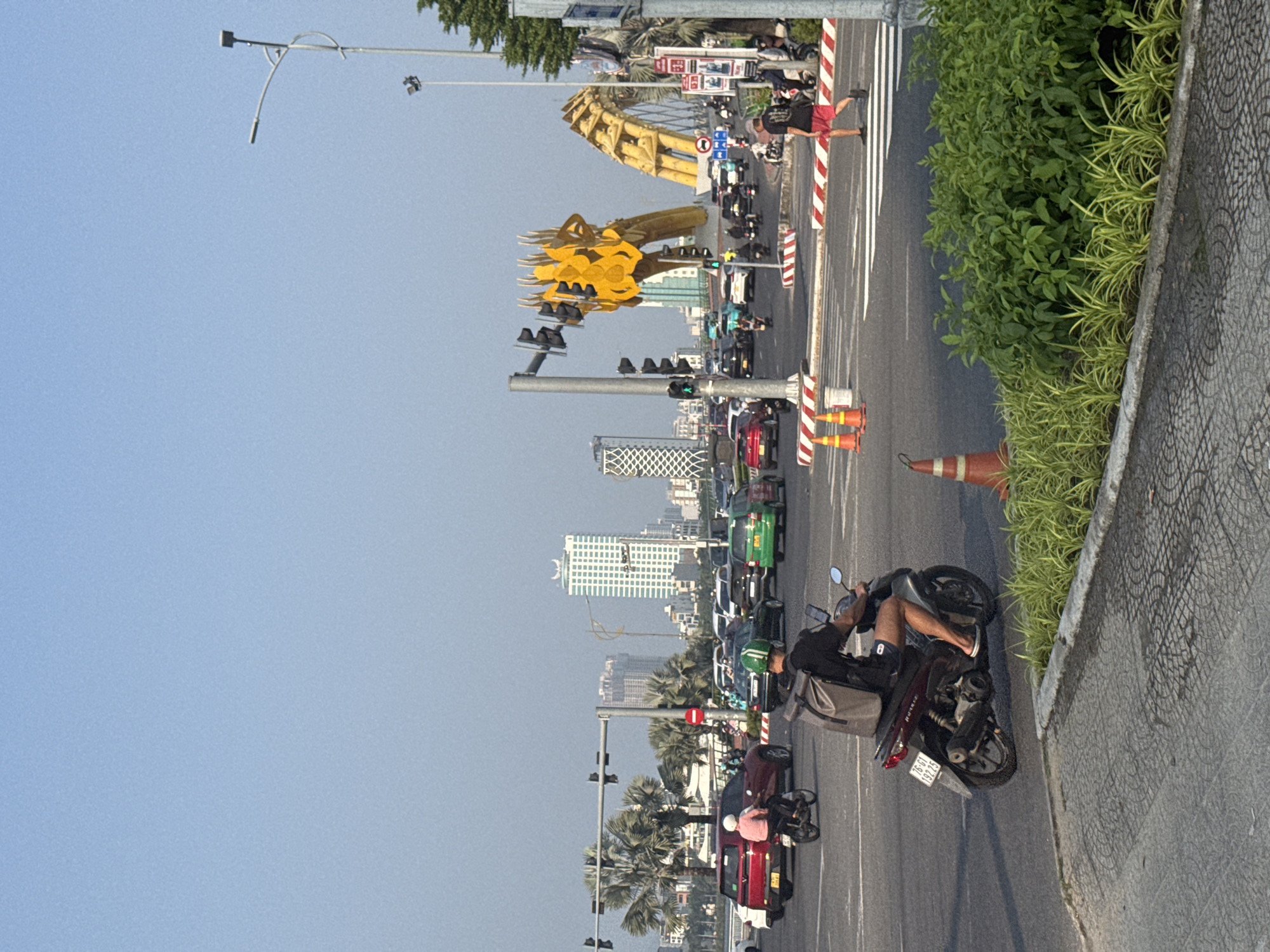 The photograph captures a street scene in Đà Nẵng, Vietnam, featuring the Dragon Bridge, a prominent landmark with a large, golden dragon sculpture. The image shows a busy street with various modes of transportation, including motorcycles, cars, and pedestrians. Traffic signals are visible along the road, indicating active traffic management. Buildings, including skyscrapers, dot the skyline in the background. A traffic cone sits on the side of the road. A man sits on a parked motorcycle on the right side of the road near a curb with foliage. The sky is clear and blue, suggesting fair weather.