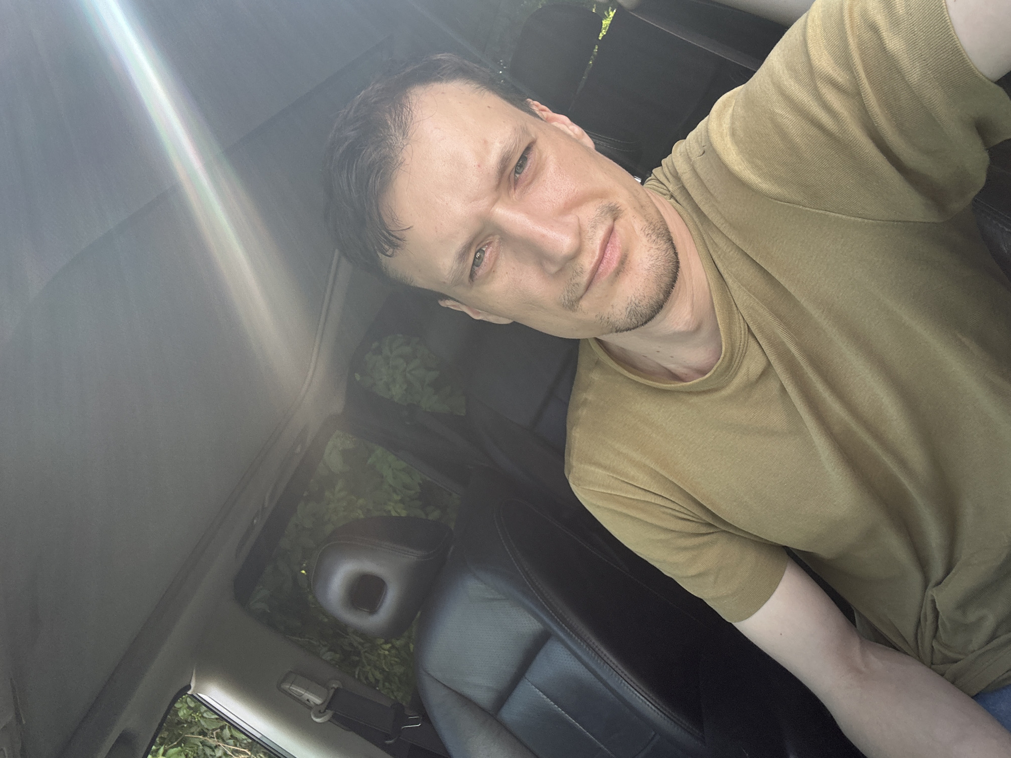 This is a selfie taken from inside a car, likely in the city of Shatava, Ukraine. The image shows a man with light skin and short brown hair, wearing a light brown t-shirt. He is sitting in the driver's seat, with the car's interior visible, including black leather seats and the headrest. Sunlight is streaming into the car, creating lens flare across the top of the image. The background outside the car appears to be lush green foliage, suggesting a location with trees and plants.