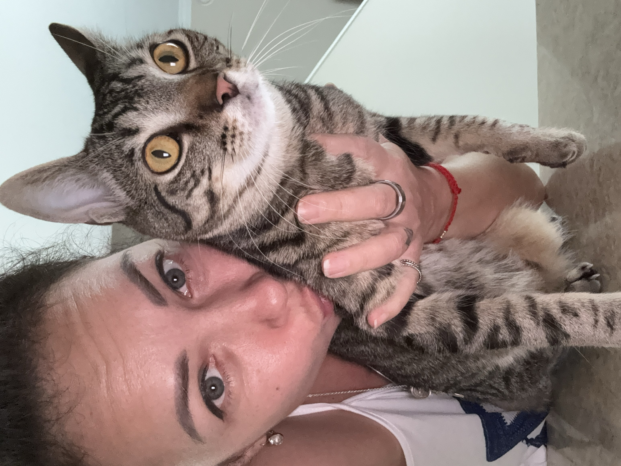 A medium shot shows a woman with dark hair pulled back, wearing a white tank top and jewelry, leaning in to kiss a tabby cat. The cat has large, wide yellow eyes and is held in the woman's left arm. The woman's right hand is around the cat's body, and her fingers are adorned with rings. A red string bracelet is visible on her wrist. The background is blurred, suggesting an indoor setting. The image appears to be a selfie, taken from a low angle.