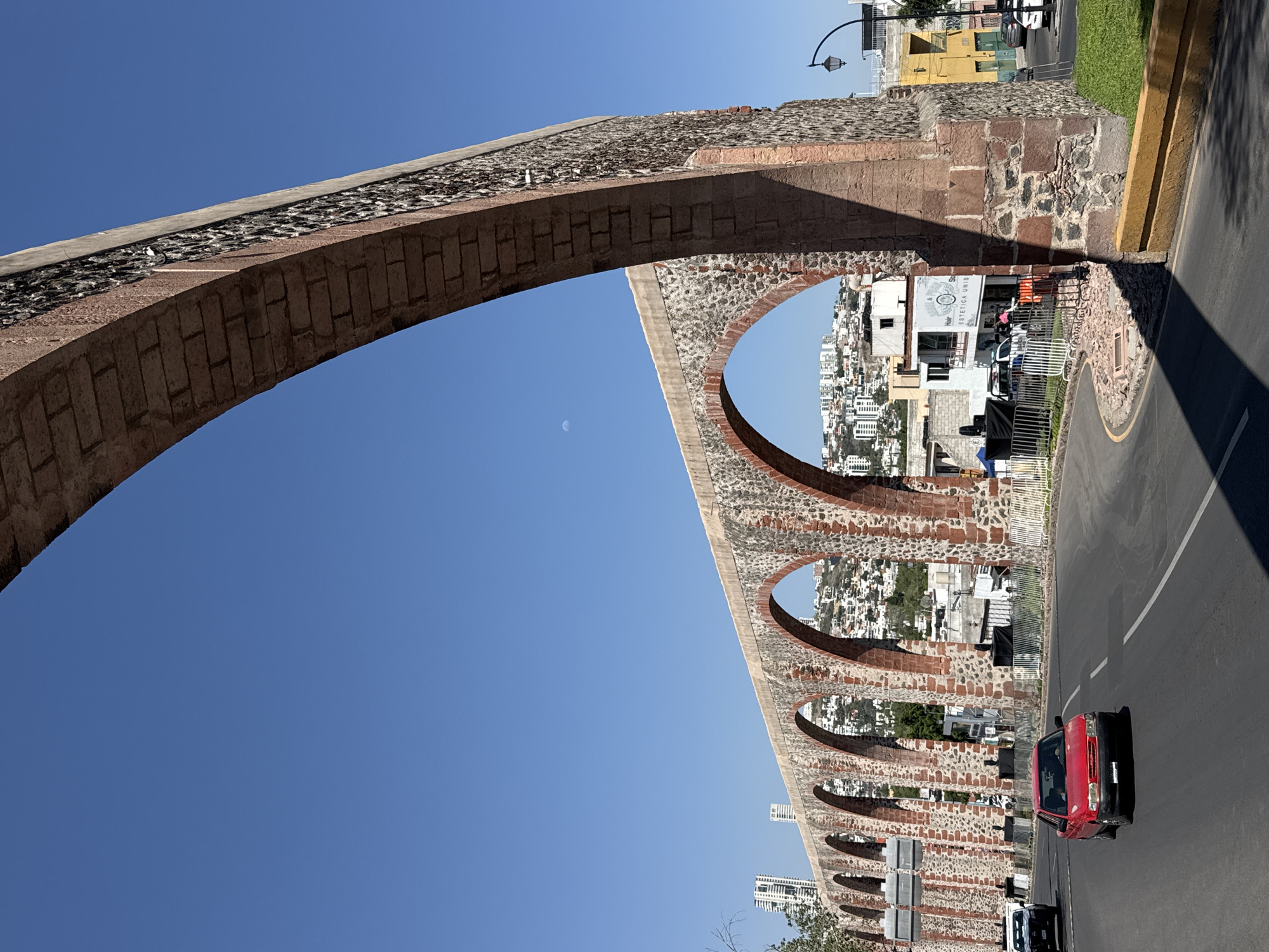 A daytime shot depicts a brick aqueduct spanning over a road in Querétaro, Mexico. The aqueduct has several arches, some of which frame views of city buildings. The sky is clear blue. A red car travels on the road under the aqueduct. The sidewalk, with its yellow curb, runs alongside the road. Buildings of varied styles and sizes are visible in the background, seen through the arches. There is a partial view of a faint crescent moon in the blue sky.