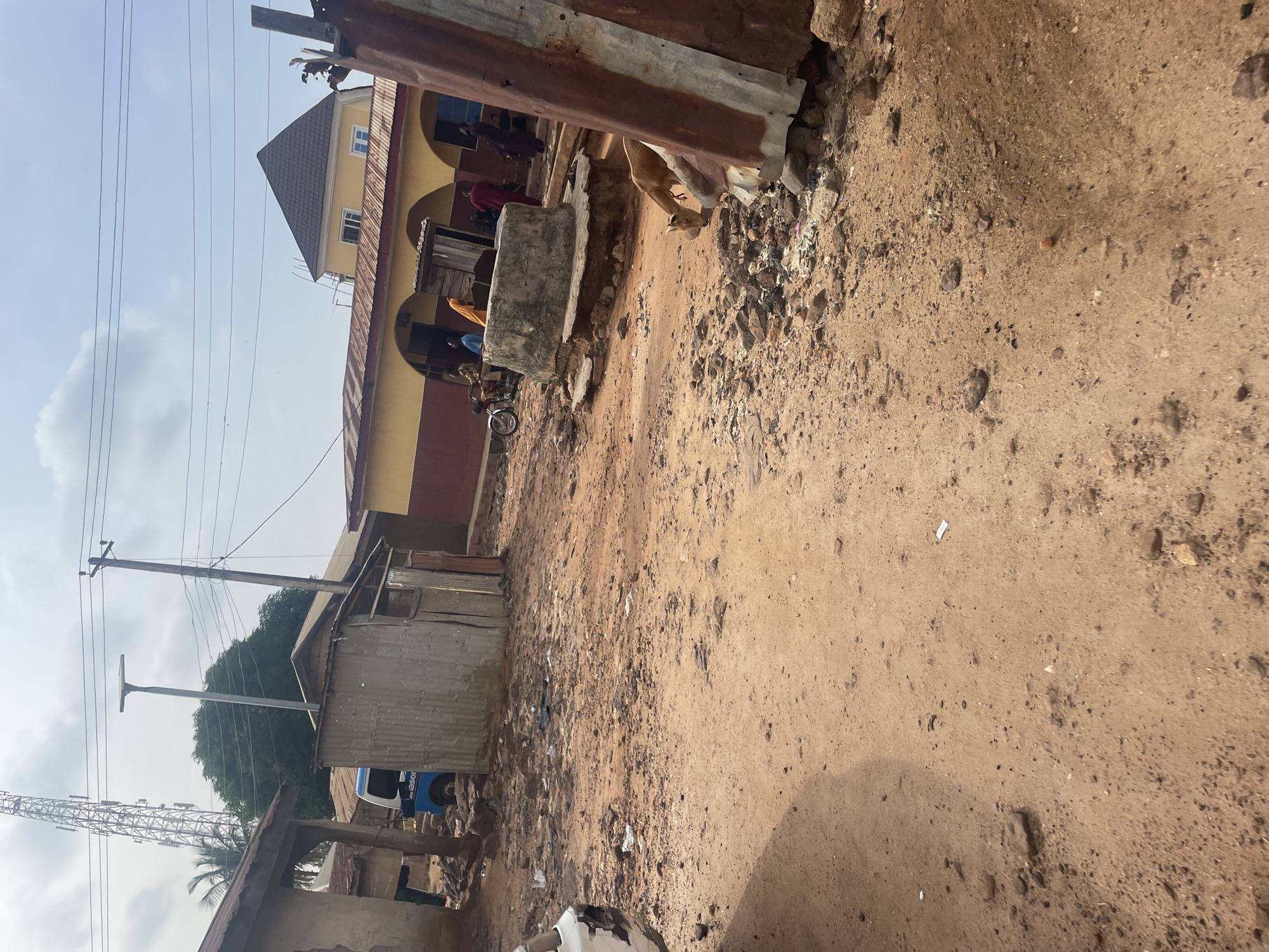 The image depicts a street scene in Lokoja, Nigeria, characterized by a dusty, unpaved road with scattered debris. In the background, a large telecommunications tower stands prominently against a cloudy sky. Several buildings line the street, including a yellow and maroon structure with arched openings and a brown, gabled roof. A corrugated metal structure, possibly a makeshift stall or shelter, is visible in the foreground. A well, constructed with concrete, is located near the center of the scene. Several people are present, some standing near the buildings and others gathered around the well. A motorcycle is parked to the left of the well. A light brown dog is seen foraging amongst the debris. A blue and white van is parked behind a corrugated metal structure. The overall impression is one of a developing urban environment with elements of traditional infrastructure and daily life.
