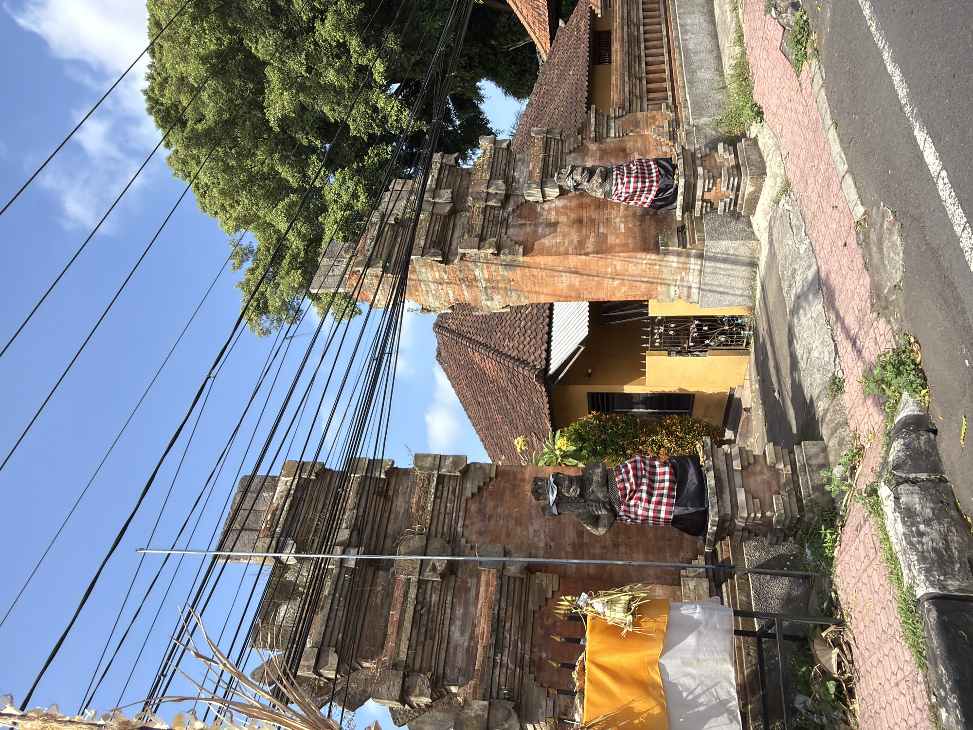 The image shows a street scene in Blahbatuh, Indonesia. In the foreground, a paved road runs diagonally from the bottom right to the bottom left of the frame. A sidewalk with red patterned pavers runs along the side of the road. On the sidewalk, there is a stone structure with a statue wearing a red and white checkered sarong. To the left of this structure, there is an ornate brick gateway with two carved figures, also wearing sarongs. A yellow cloth is draped on the left side of the gateway, adorned with offerings. Behind the gateway, a house with a red tile roof and yellow walls is visible. A large tree with green foliage dominates the upper right portion of the image. Numerous electrical wires are strung across the sky, obscuring a clear view of the blue sky. The overall impression is of a tropical, urban environment.