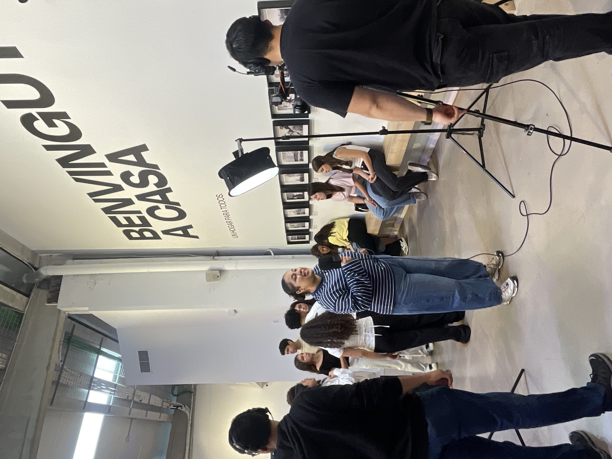 The image depicts a group of people standing in a line in what appears to be an indoor space. They are facing towards the right of the frame, possibly participating in a recording or filming session. Two individuals are operating camera equipment, each wearing headphones and focused on the subjects. Lighting equipment is also present, suggesting a professional setup. The environment features a wall with the inscription "BENVINGUT A CASA" and "UN HOGAR PARA TODOS", possibly indicating a branding or campaign message. The architectural style and the presence of the Catalan phrase suggest that the location could be in Barcelona, Spain. The setting seems like a studio or a large open area used for media production.
