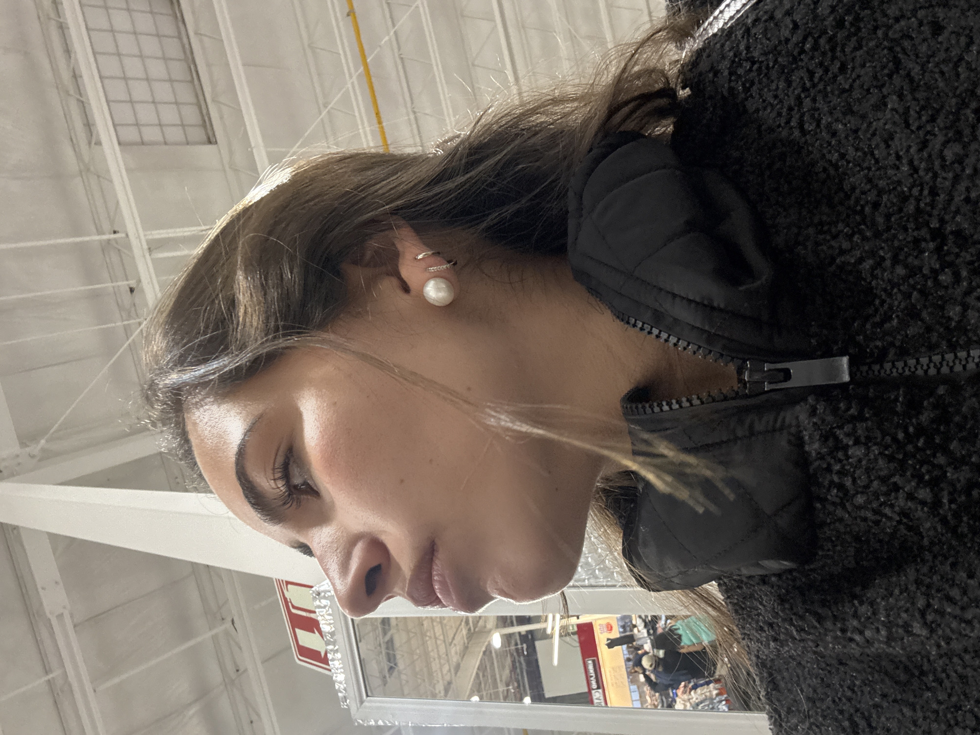 A woman is positioned inside what appears to be a retail environment, possibly a Costco store, given the architecture of the ceiling. She is wearing a black, quilted jacket with a textured, dark-colored coat over it and pearl earrings. A sign with a red arrow points to the left. The location is potentially Puebla, Mexico.