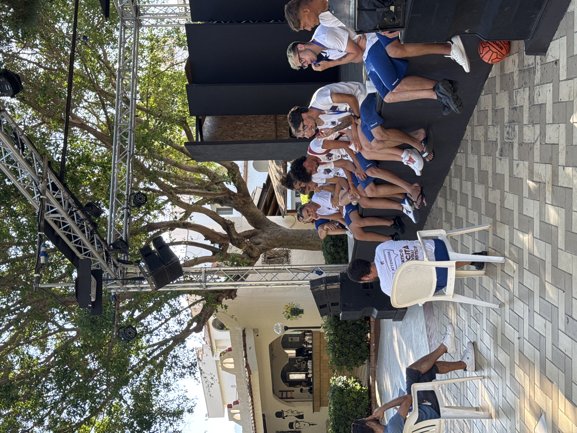 In Isola delle Femmine, Italy, a group of young people are seated on a low stage outdoors. The setting appears to be a venue with a tiled floor and a large tree providing shade. A white building with arched openings is visible in the background, suggesting a restaurant or public space.
The individuals are mostly wearing white t-shirts and blue shorts, some with what appear to be lanyards around their necks. They are casually dressed and some are engaged in conversation or looking at their phones. A few are seated in white plastic chairs in the foreground, observing the group on the stage.
The stage is equipped with sound equipment, including speakers. A red soccer ball rests on the floor near the speakers. The overall atmosphere is relaxed and informal, possibly during a break or presentation at an event. The lighting suggests it is daytime.