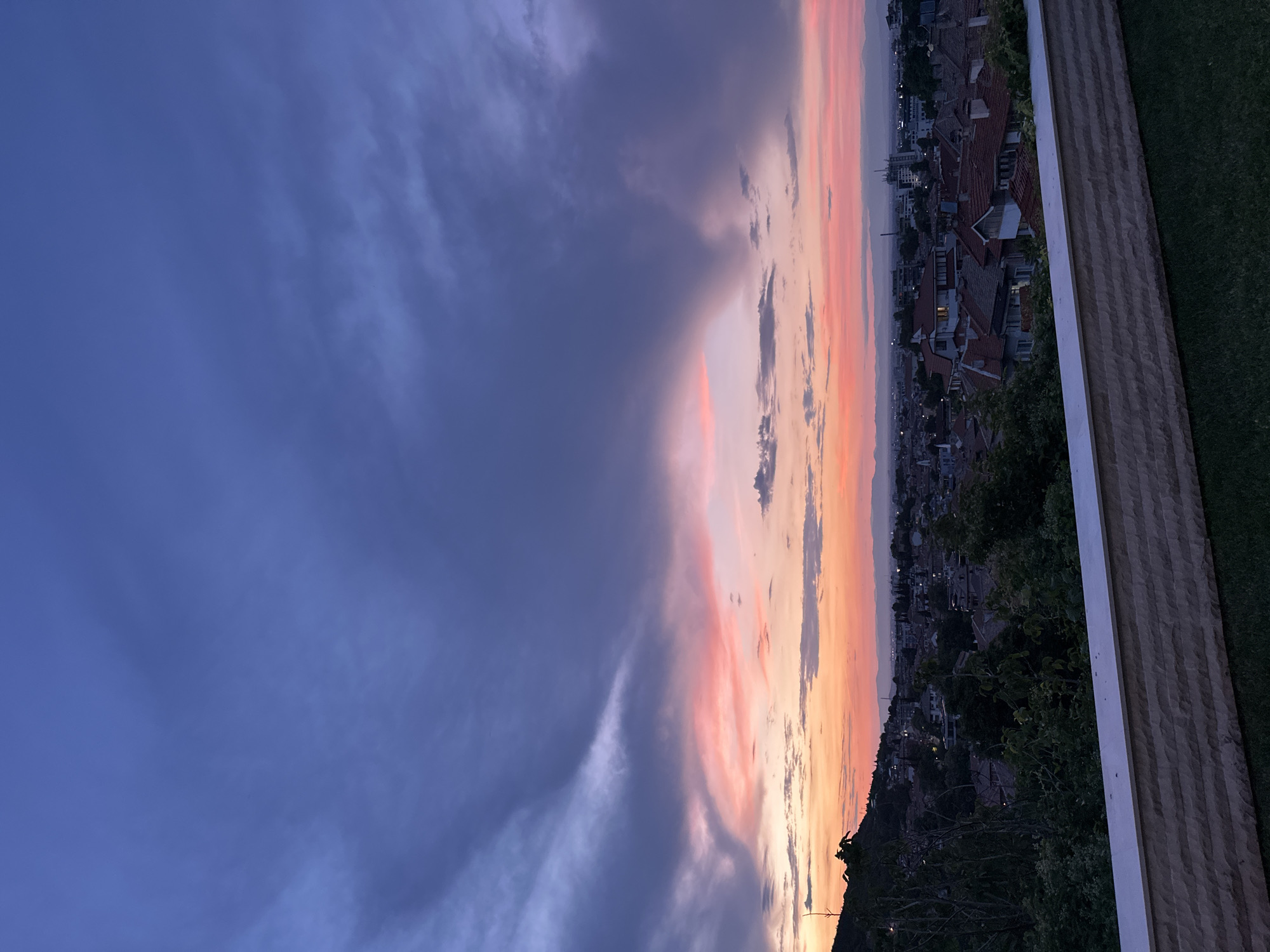 The image depicts a twilight view over the city of Asenovgrad, Bulgaria. In the foreground, a low, light-colored stone wall with a textured surface runs horizontally across the bottom of the frame, with a patch of manicured green grass visible below it. Beyond the wall, a dense canopy of dark green trees obscures much of the lower portion of the cityscape, with glimpses of red-tiled rooftops and some white-walled buildings peeking through.
The middle ground shows a sprawling urban landscape with a variety of building architectures, dominated by the warm tones of terracotta roofs. The city extends towards a distant horizon marked by a range of hazy mountains. The sky above is a dramatic canvas of dusk colors. Dark, heavy clouds with shades of deep blue and purple dominate the upper portion of the image, creating a sense of depth and movement. Lower in the sky, a band of soft pink, orange, and yellow hues illuminates the horizon, indicating the setting sun. The overall atmosphere is serene and captures the transition from day to night over the city. No people or specific actions are visible in the image.