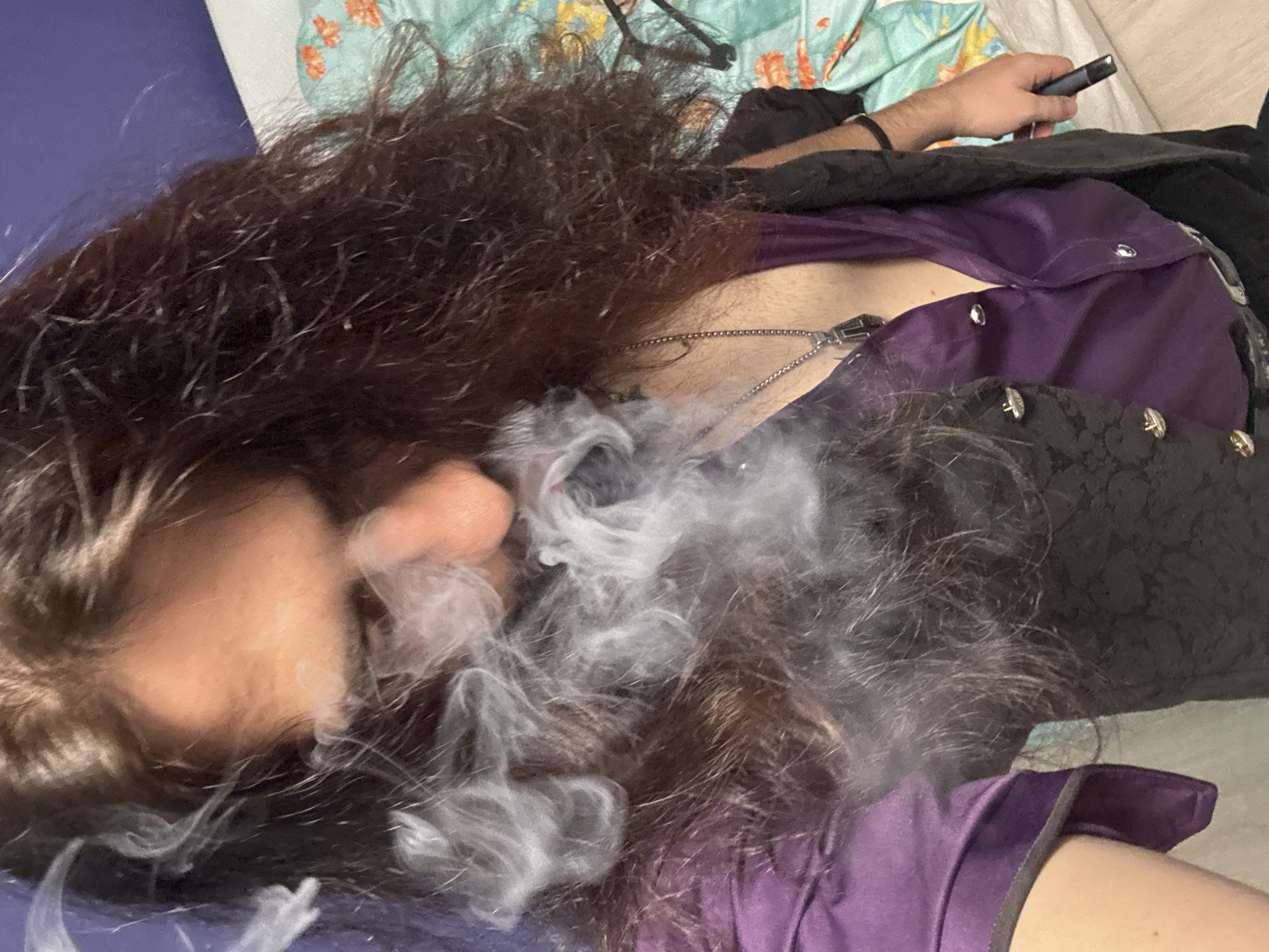 The image shows a person with long, dark, curly hair. They are wearing a purple button-up shirt and a dark vest. A chain with a pendant is visible around their neck. The person appears to be exhaling smoke or vapor, which obscures part of their face. They are holding a dark electronic device in their right hand. The background includes a bed with a patterned comforter and a blue wall. The scene appears to be indoors.