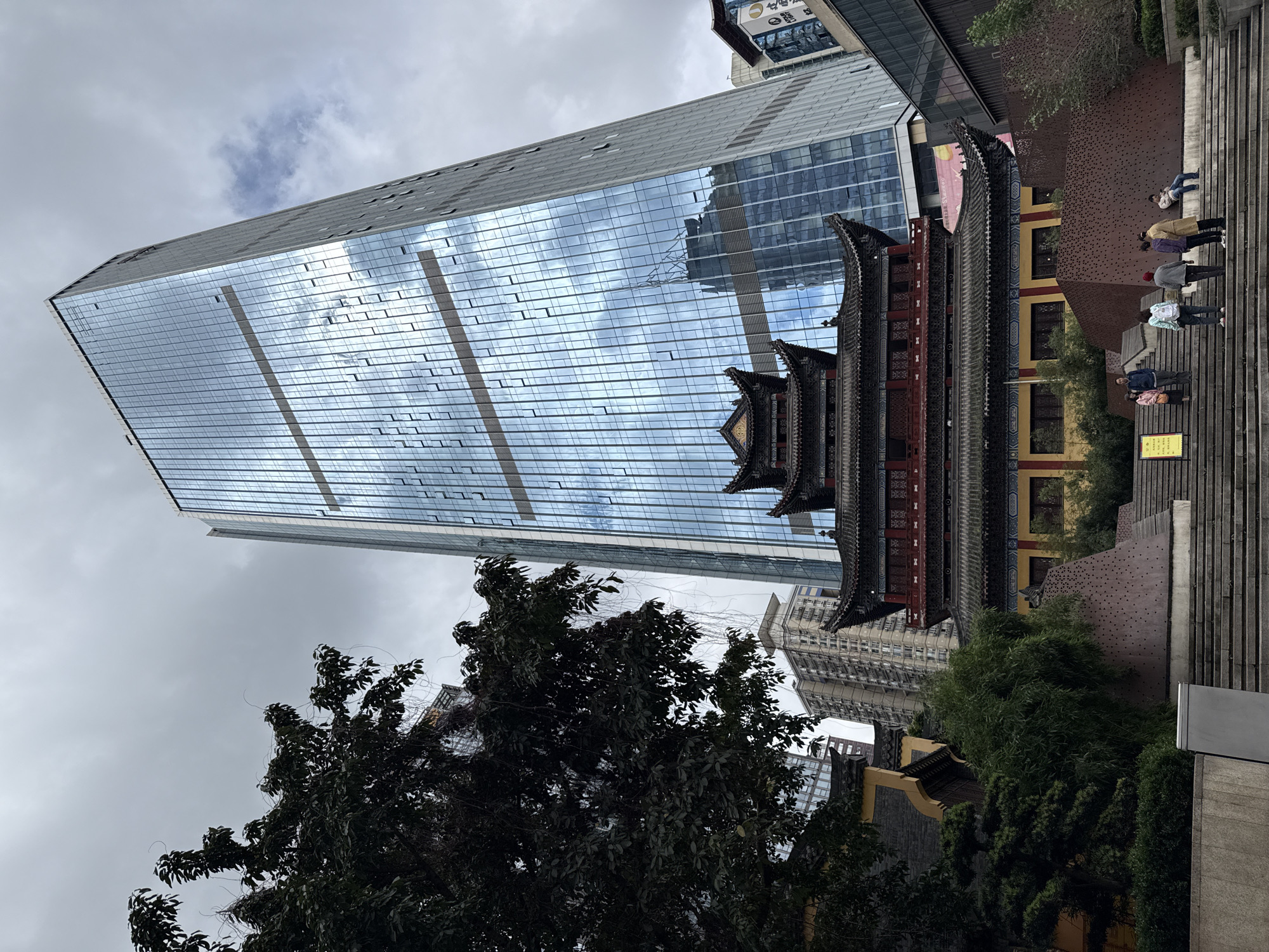 The image depicts a modern glass skyscraper situated adjacent to a traditional Chinese pagoda structure, indicative of the urban landscape of Yuzhong District, China. The skyscraper's reflective facade mirrors the cloudy sky. In front of the buildings, green trees obscure the bottom portion of the frame. To the right of the pagoda, several people are walking up stairs. A sign is visible on the far right of the image.