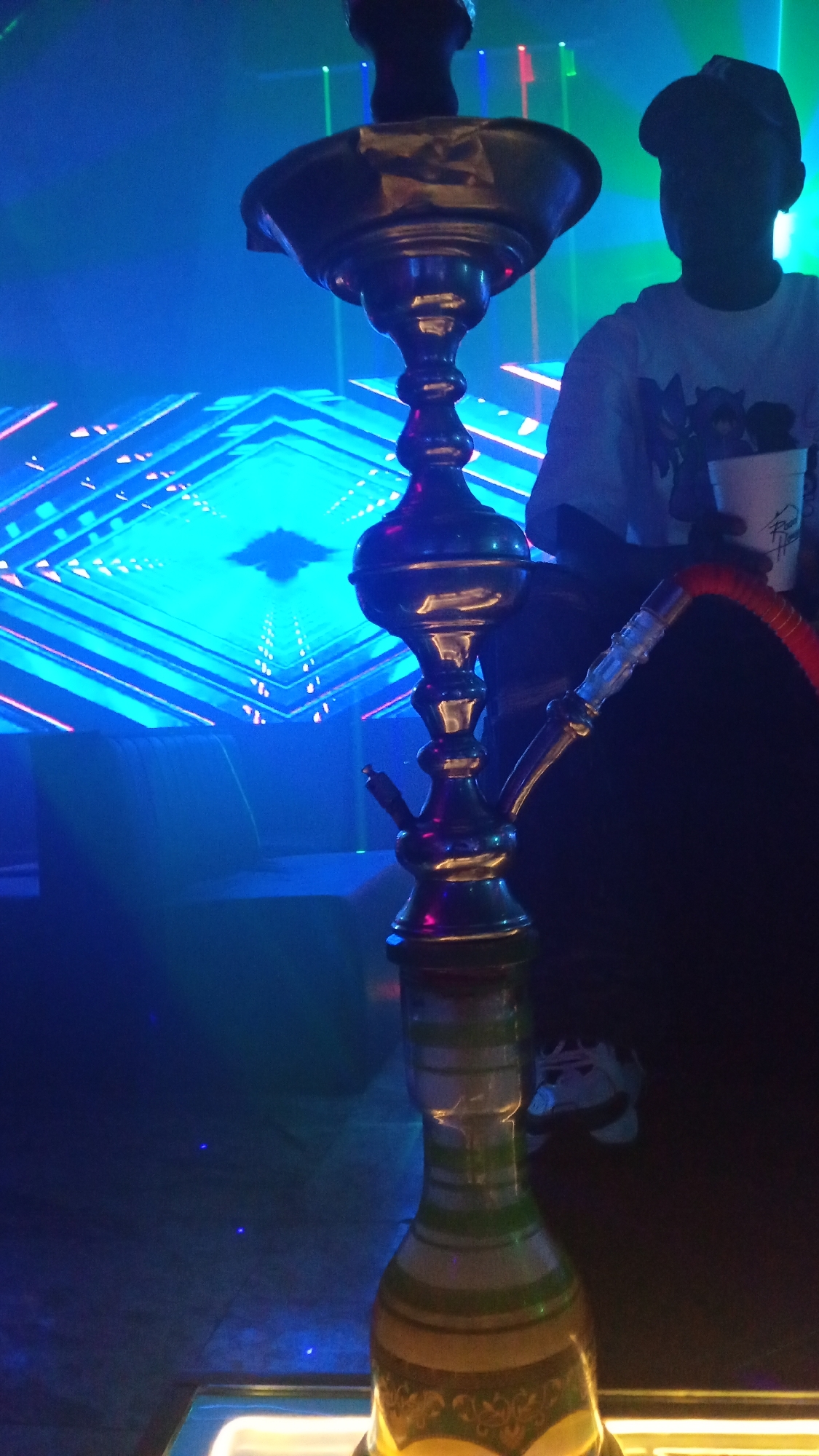 A person in a dark room with a hookah pipe in the foreground. The person is wearing a white t-shirt and a cap, holding a white cup. The background is illuminated by blue and green lights, with a geometric pattern projected onto a wall. The hookah pipe is made of metal and glass, with a red hose. The overall atmosphere is dark and moody, with a focus on the hookah pipe. The scene could be a lounge or a club in Howayi, Nigeria, where people gather to socialize and smoke hookah.