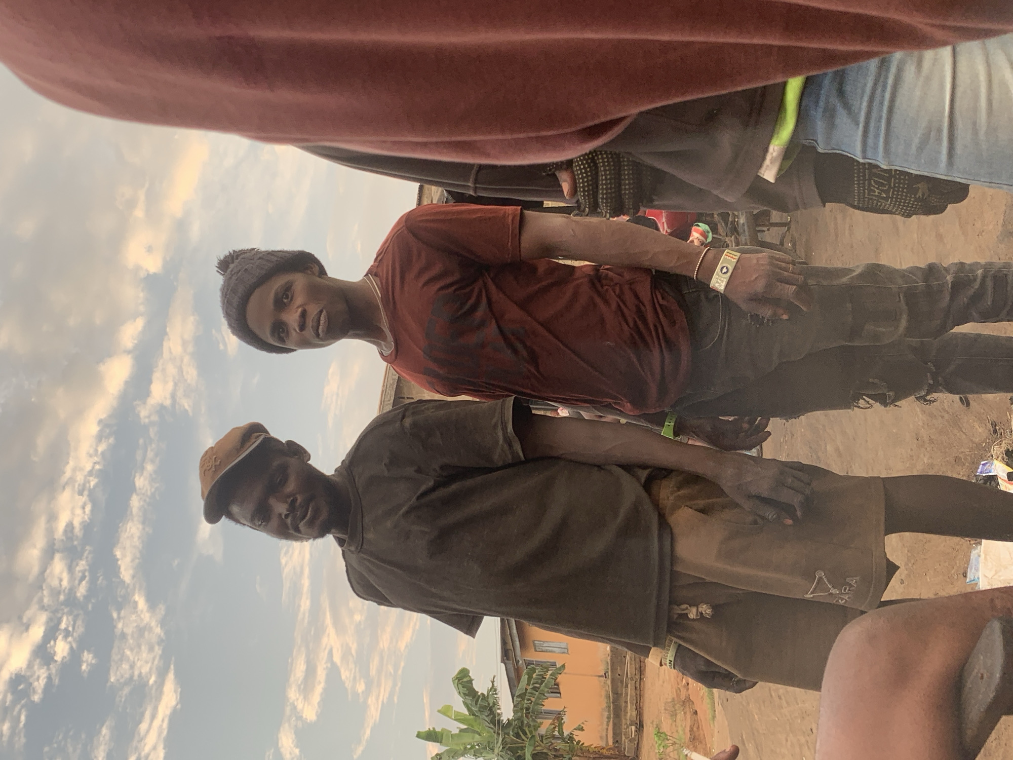 The image depicts an outdoor scene in the city of Shagamu, Nigeria. Two men are the primary subjects, standing close together. The man on the left wears a brown baseball cap, a dark t-shirt, and brown shorts. The man on the right is wearing a dark beanie, a reddish-brown t-shirt, and ripped dark pants. A portion of a third person, wearing a burgundy garment, is visible in the foreground on the right. The background shows a building with orange walls and a banana tree, suggesting a residential or semi-urban environment. The ground is dirt, and there are some scattered items, possibly debris or merchandise, on the ground. The sky above is partly cloudy, with the light suggesting it might be late afternoon or early evening.