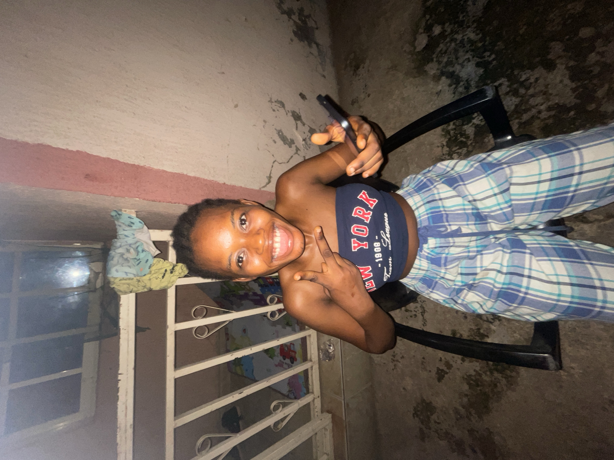 A young woman is sitting on a black chair outside a building in Lokoja, Nigeria. She is holding a smartphone, and giving a peace sign to the camera. She wears a dark-colored tank top with "New York" written on it, and light blue and white plaid pants. The background consists of a white wall with a red painted strip, a window, and a metal gate. The ground is made of cement and has a soiled appearance.