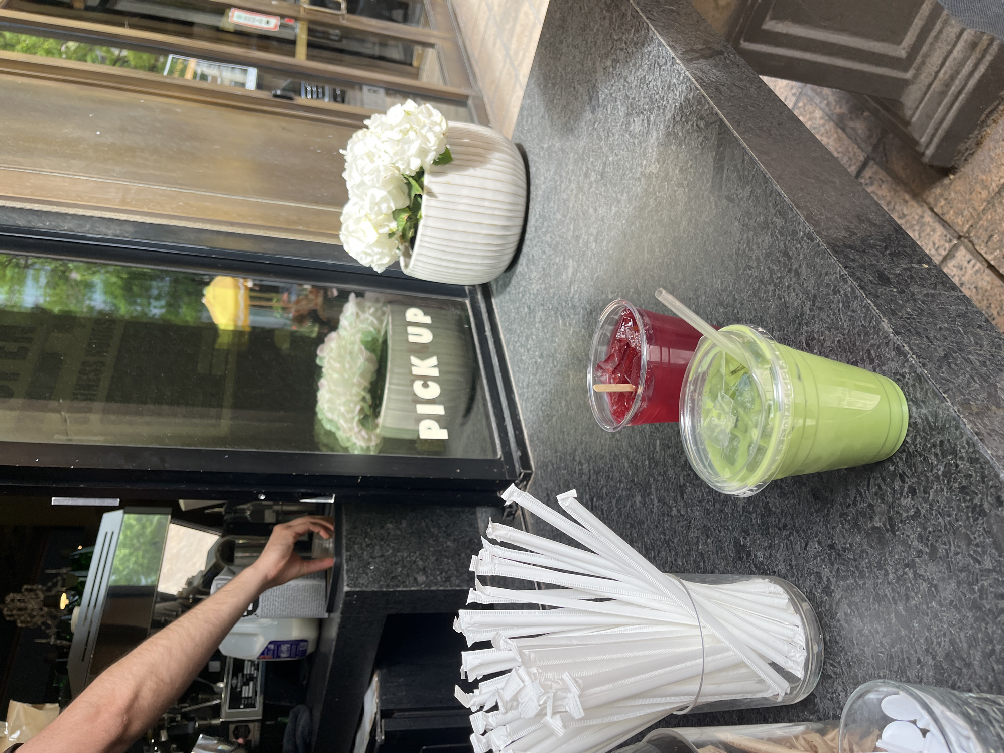 This image shows a view of a cafe counter with several items, possibly a pick-up window, in Austin, United States of America.
On the counter are two beverages in clear plastic cups: one with a red drink and another with a green drink, each with a straw. There is also a glass jar filled with white paper-wrapped straws. Next to the pick-up window, there's a white flower pot filled with white flowers.
A person's arm is visible reaching into what appears to be the cafe's interior, suggesting someone is working behind the counter. The reflection in the pick-up window shows the outside surroundings, including greenery and what could be street activity. The counter itself is made of a dark, speckled material.