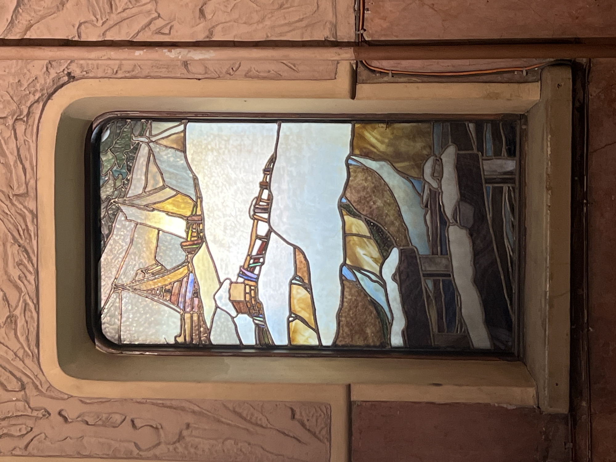 This image features a rectangular stained glass window embedded within a textured wall. The stained glass artwork presents an abstract or semi-abstract composition, utilizing various geometric and organic shapes. The color palette primarily consists of muted and warm tones, including whites, creams, yellows, browns, greens, and touches of blue and red. The arrangement of glass pieces evokes a sense of a rugged landscape, possibly depicting rocky formations, perhaps a waterfall or flowing water in the blue sections, and distant structures. The glass itself appears textured, allowing light to diffuse through it.
The stained glass window is framed by a smooth, light beige or cream-colored recess with rounded corners, which contrasts with the rougher, relief-carved texture of the surrounding wall. The wall material is a reddish-brown, suggesting stone or heavily textured plaster with an ancient or rustic aesthetic. Exposed orange and brown electrical wires are visible along the top right edge of the window frame and wall. The stained glass is illuminated from behind, suggesting either natural light from an exterior source or deliberate backlighting. No people are visible, and no specific activity is taking place within the static scene. There is no discernible text in the image. The overall impression is one of intricate architectural detail, characteristic of early 20th-century design often found in cities like Saint Petersburg.
