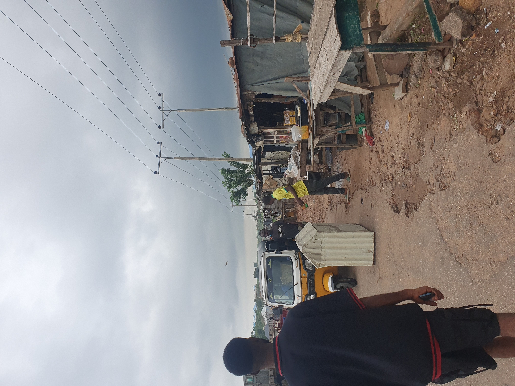 This is a full-body, eye-level shot captured in Lokoja, Nigeria, focusing on a street scene with several individuals and a tuk-tuk. The sky is overcast, suggesting a cloudy or potentially rainy day. In the foreground, a man wearing a black t-shirt with red trim and black shorts is walking away from the camera, holding a dark object in his right hand. To his left, a yellow and white tuk-tuk is parked on a dirt road. Behind the tuk-tuk, two men are visible; one in a dark t-shirt and another in a bright yellow t-shirt. The background depicts a row of makeshift stalls or shops, constructed from various materials like corrugated metal and tarpaulin, lining the dirt road. These stalls appear to be selling goods, with some items visible on display. Overhead, power lines are strung between utility poles, a common sight in urban and semi-urban areas. The ground is uneven and appears to be a mix of dirt, gravel, and some debris. The overall impression is of a bustling, informal marketplace in a developing urban setting.