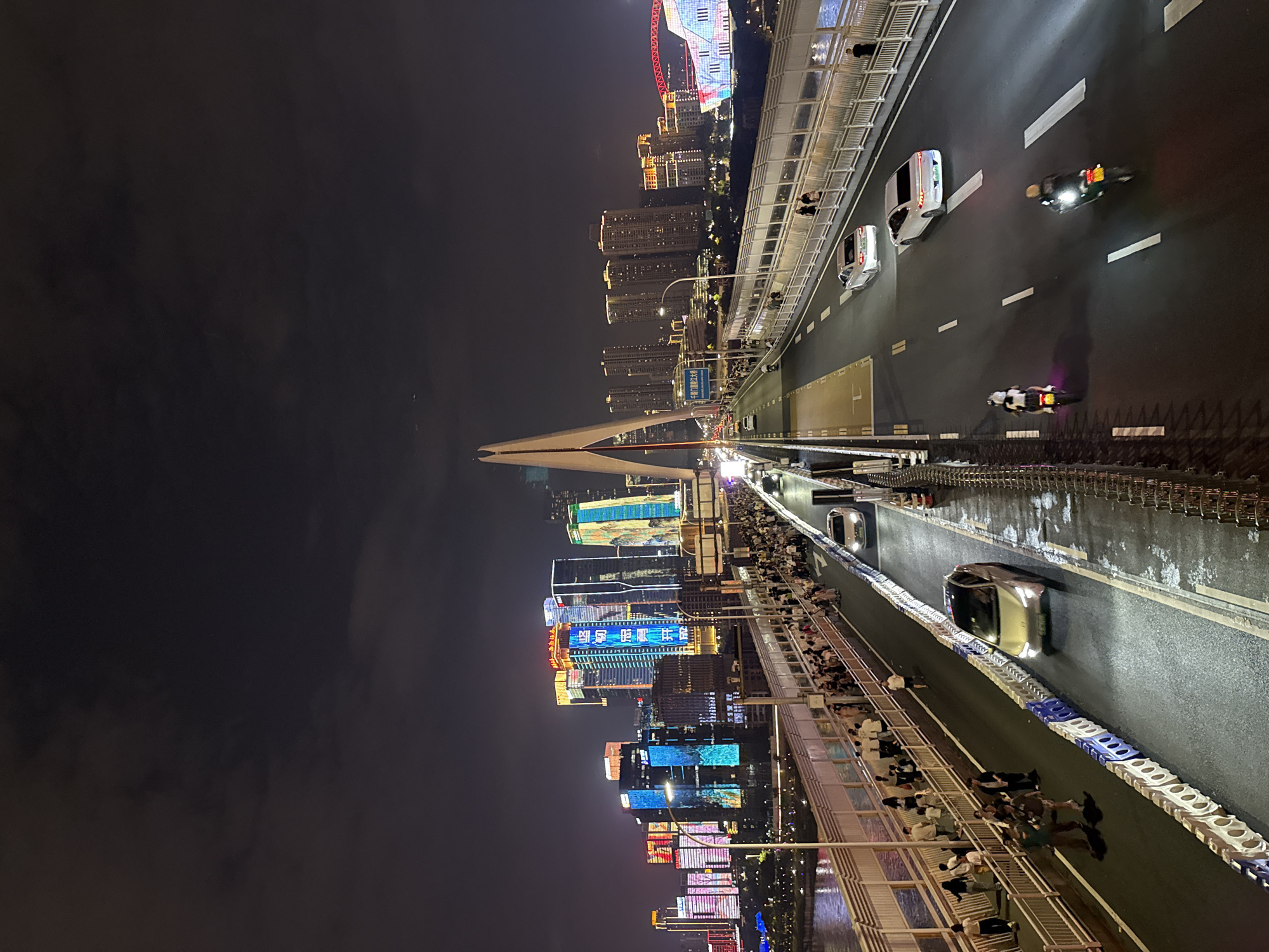 The image captures a nighttime view of a modern cityscape with a focus on a bridge, likely in Yuzhong District, China, given the architectural style and illuminated skyscrapers. The bridge is multi-layered, with a pedestrian walkway on the left and a vehicular roadway on the right. The pedestrian walkway is densely populated with people strolling and observing the city. Several cars and motorcycles are traveling on the roadway. The background is dominated by tall buildings adorned with colorful neon lights and digital displays, creating a vibrant urban panorama. The sky is dark, suggesting it is nighttime, with some clouds present. A prominent, uniquely shaped white pylon structure is visible on the bridge.