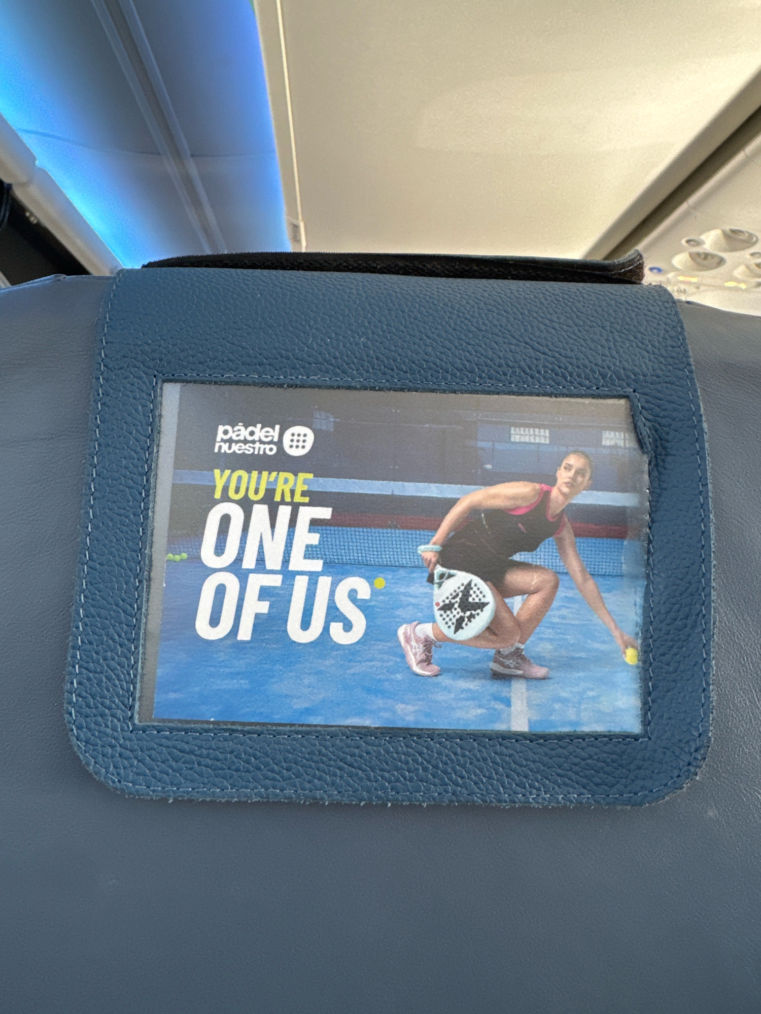 The image shows a close-up of a seatback pocket on an airplane, likely in flight. A promotional image for "Padel Nuestro" is displayed within the pocket. The image features a woman playing padel tennis on a blue court, with the text "YOU'RE ONE OF US" prominently displayed. The seatback is made of dark blue faux leather. The overhead cabin lighting, emitting a cool blue hue, is visible at the top of the frame. The context of "Padel Nuestro" and the generic airplane interior suggest a potential advertisement displayed during travel, possibly targeting individuals with an interest in sports or recreational activities. There is no specific indication of the city of Palma de Mallorca, Spain.