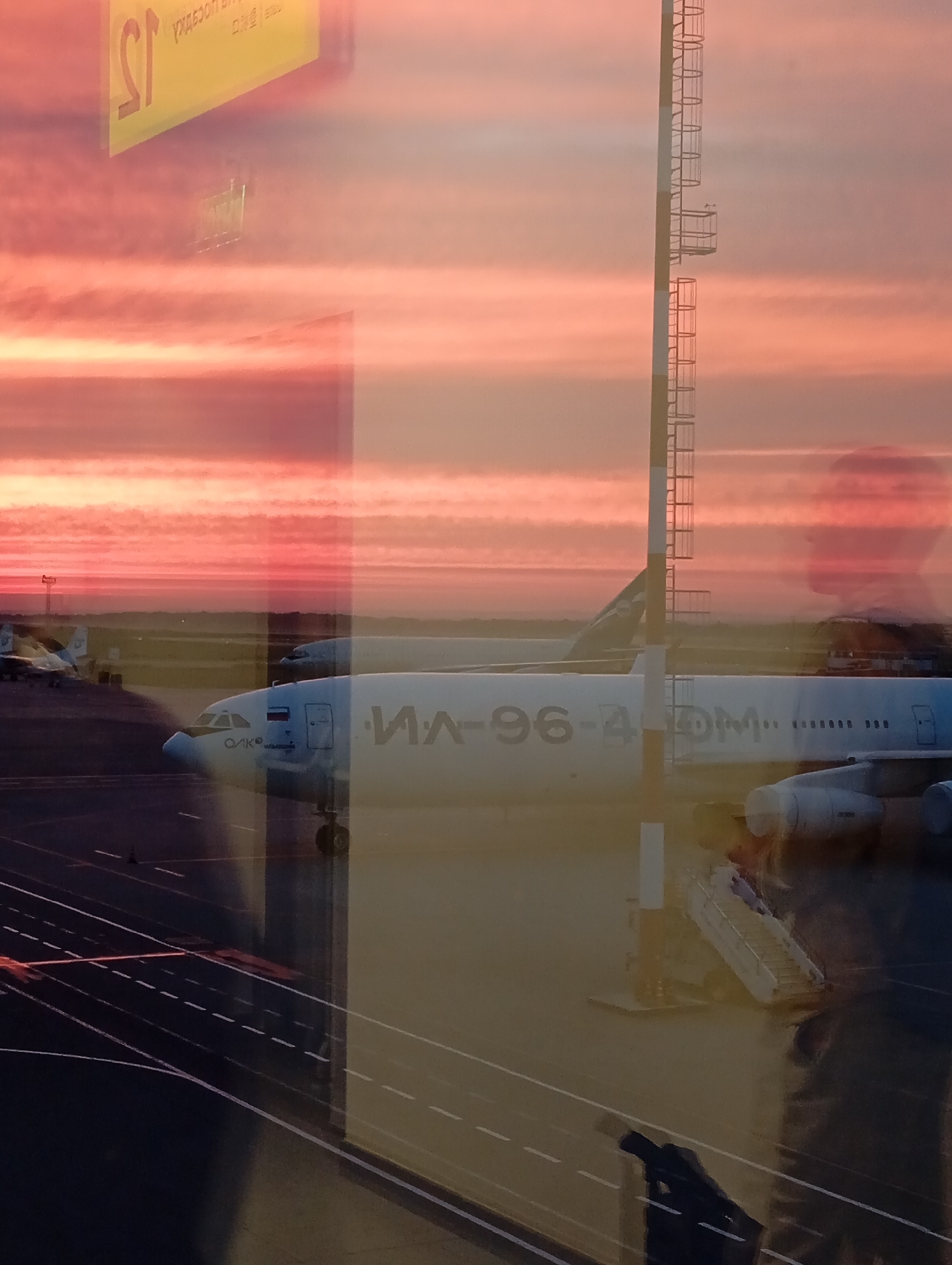 This image captures a reflection of an airport tarmac during a sunset, likely at Khabarovsk Novy Airport in Khabarovsk, Russia. The sky is a vibrant gradient of pink and orange, with thin clouds streaking horizontally. In the foreground, a large airplane, identified by the Cyrillic text "ИЛ-96-400" on its fuselage, is parked on the tarmac. The plane is white with Russian flag insignias on its tail.
Multiple reflections are visible on the surface from which the photo was taken, possibly a window. These reflections include a silhouette of a person's head and shoulders, a yellow sign with Cyrillic text (partially visible as "SY"), and part of a tall, striped structure resembling a control tower or equipment stand with ladders. Other airplanes are faintly visible in the background. The tarmac is marked with white lines, and a set of mobile stairs is positioned near the airplane's entrance. The overall scene depicts a moment of transition at an airport as day turns to night, with the dramatic sunset colors creating a striking visual contrast with the industrial elements of the airport.