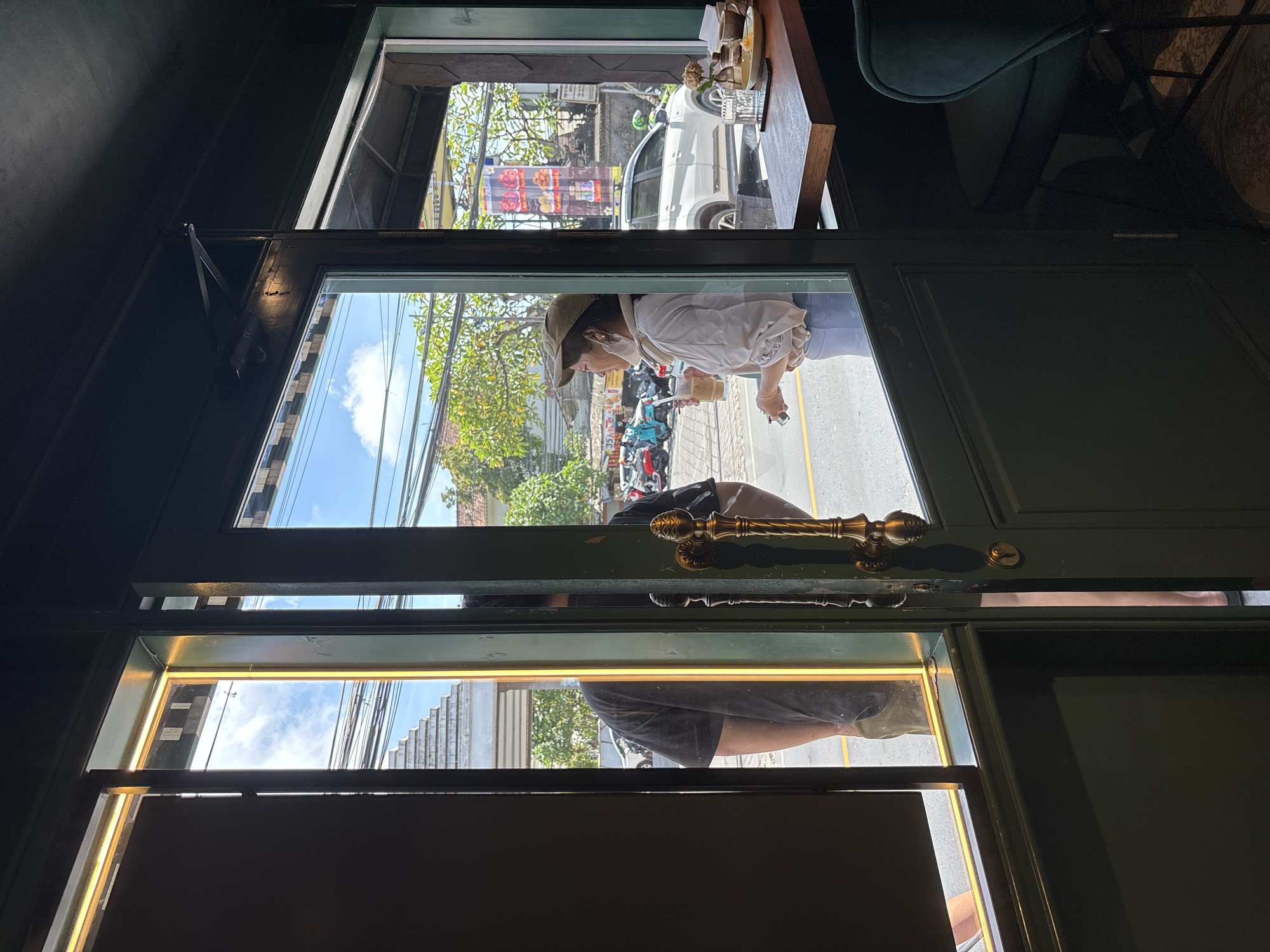 The image shows a partial view through a glass door, likely of a commercial establishment in Ungasan, Indonesia. Through the upper pane, a woman is visible in the street, holding a beverage and a cell phone. She is wearing a cap, a white shirt, and light-colored pants. Behind her, there are parked motorbikes and trees lining the street. A white car and storefronts are visible further in the background. Through the lower pane, another person is partially visible, wearing a dark t-shirt. The door has a brass handle and locking mechanism. Inside, a table with items on it and a green velvet chair can be seen.
