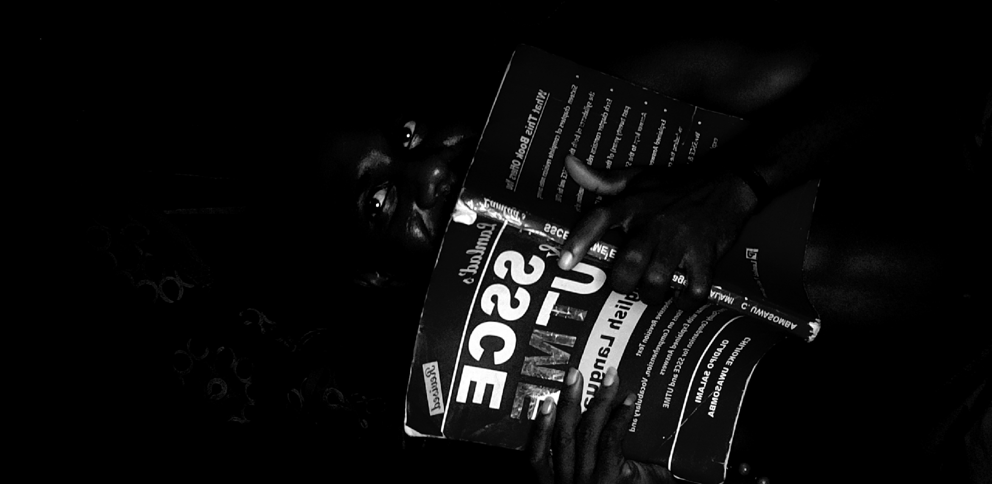 A close-up, low-angle, black-and-white photograph shows a person holding two copies of "Family's Choice Super Daily 22CE" newspapers. Only part of the person's face is visible, including their eyes and nose. The newspapers are stacked and angled toward the viewer. There are some barely discernible shapes on the dark, cluttered background. It is not possible to determine the location or the city of Narayi, Nigeria, from the image.