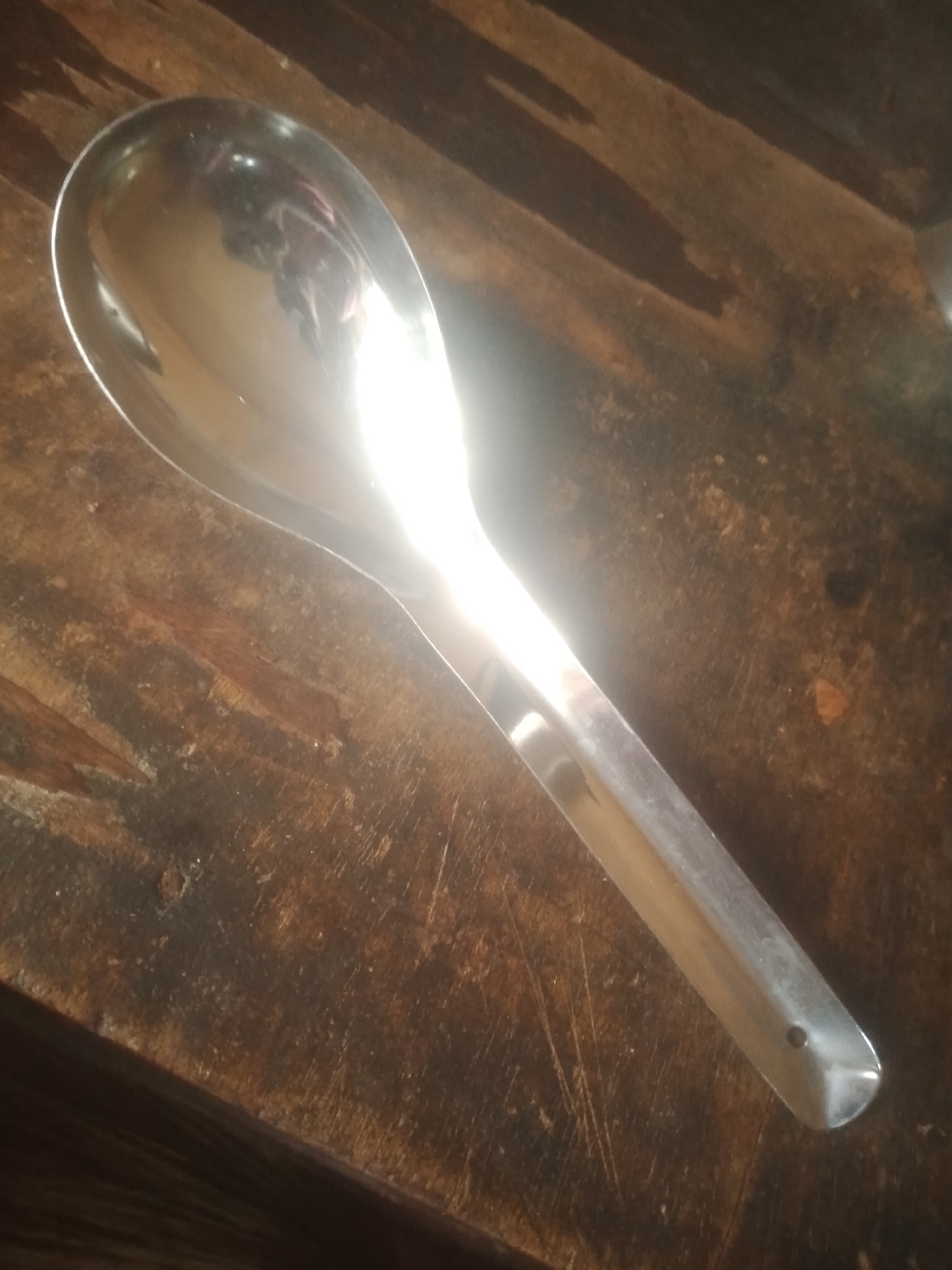 A large, shiny metal ladle rests diagonally on a rustic wooden surface. The ladle is positioned with the scoop end towards the upper left of the frame, reflecting bright light and indistinct shapes. The wooden surface appears to be a table or countertop with visible grain, scratches, and variations in color, ranging from dark brown to lighter reddish-brown hues. The lighting creates strong highlights on the ladle, obscuring some of the details of the surface beneath it. The background is out of focus, further emphasizing the ladle and the immediate wooden surface. There are no people, animals, or other objects clearly visible in the image. The scene appears to be indoors, given the lighting and the nature of the objects. This image is not located in the city of Jauro Yinu, Nigeria.