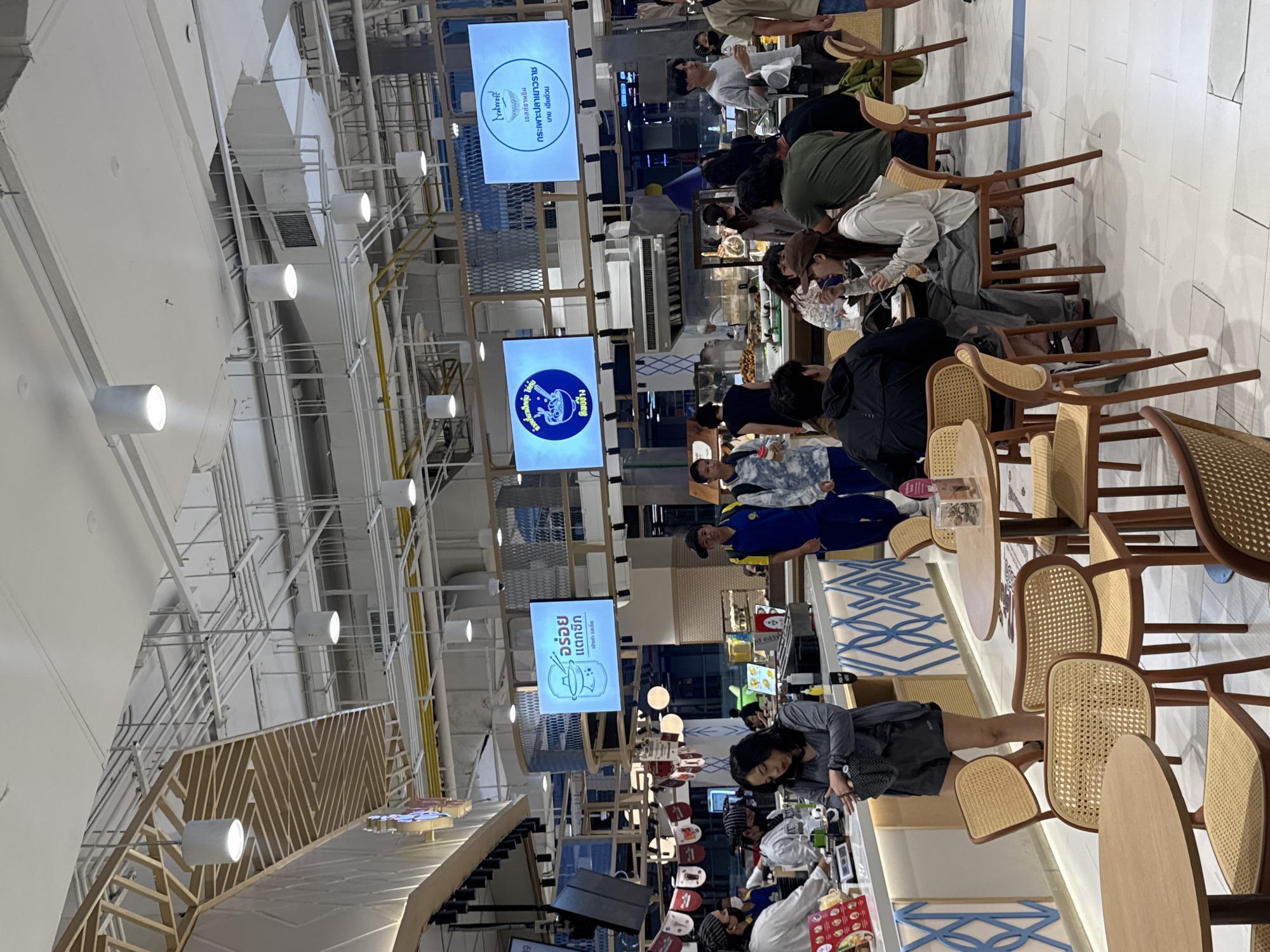 The image depicts a bustling indoor food court, likely a modern establishment with a light and airy design. In the foreground, several patrons are seated at round wooden tables, some engaged in eating, others in conversation. The chairs are of a modern design with woven cane seats and backs and wooden frames. The floor is tiled in a white checkered pattern. In the midground, a food counter is visible, with staff members serving customers. Behind the counter, various food displays and cooking equipment are present. Large digital screens are mounted above the counter, displaying menu items or advertisements in Thai script. The ceiling is a complex network of lighting fixtures, ventilation ducts, and structural beams, characteristic of a large commercial space. The overall atmosphere appears to be one of casual dining and commercial activity. Based on the signage in Thai, the location is in the city of Nong Pa Khrang, Thailand.