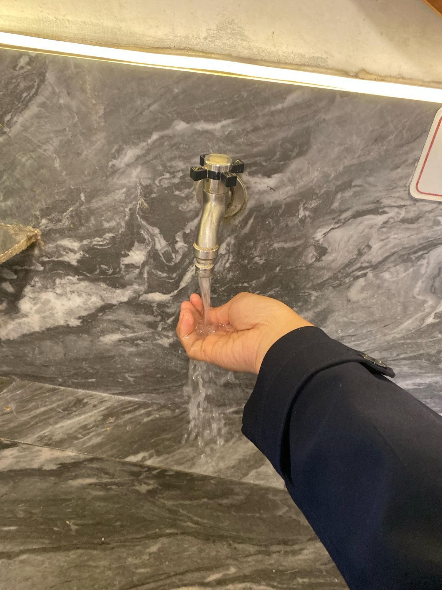 A person's hand, with a navy blue sleeve visible, is held under a silver faucet as water streams into their cupped palm. The background is a grey, marbled wall. The scene is lit by an overhead light fixture. The location is not specified.