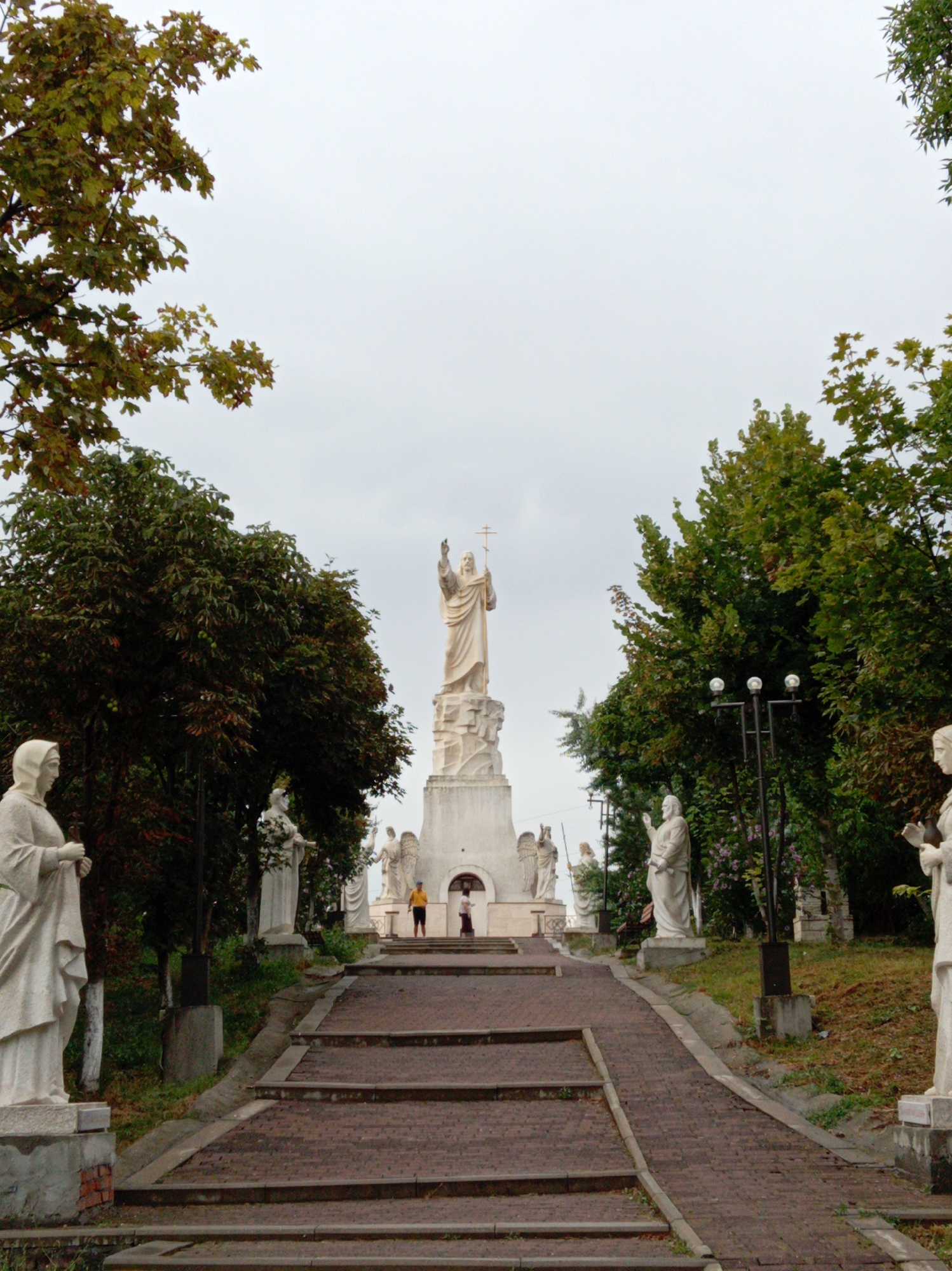 The image depicts a large outdoor sculptural ensemble, likely a memorial or religious site, with multiple statues of human figures. The main focal point is a tall statue of Jesus Christ at the top of a hill, with his right hand raised. A pathway of brick steps leads up the hill, flanked by smaller statues on either side, some appearing to be angels or saints. Lush green trees and foliage surround the area, with some trees showing autumn colors. The sky is overcast and gray. In the foreground, on the left, is a statue of a robed woman. On the right, a statue of a robed man is visible. Further up the pathway, two figures are seen standing near a building. The entire scene is set in an outdoor environment with a clear, overcast sky. This location is the "Mountain of Forgiveness" in Yessentuki, Russia.