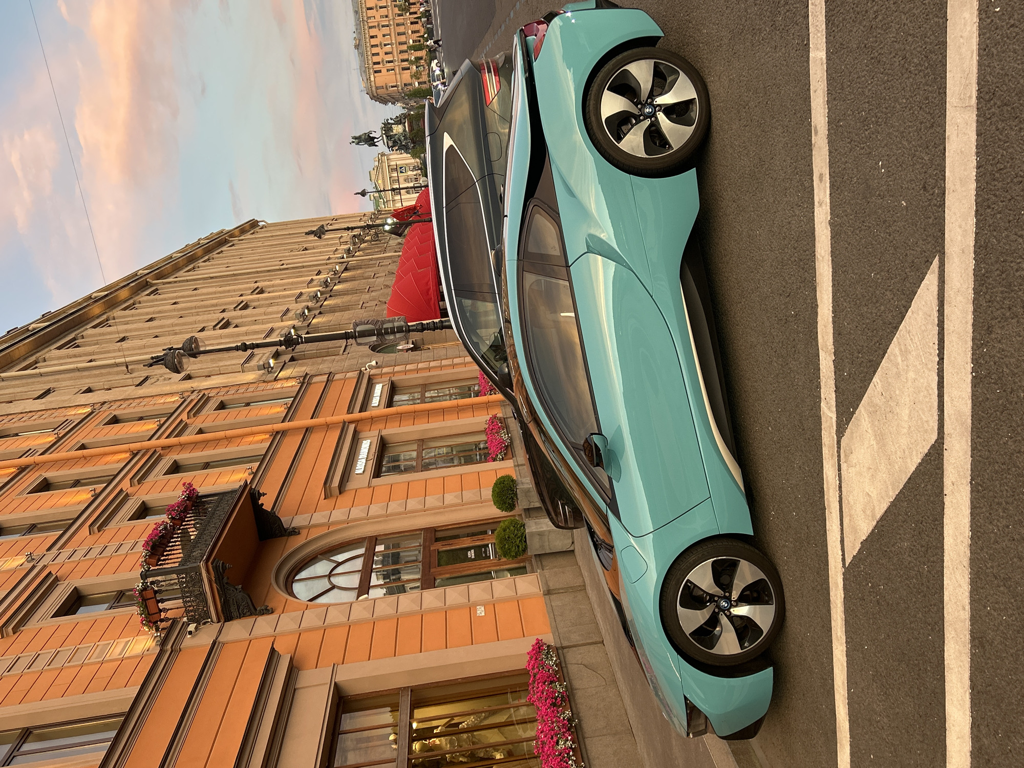 An eye-level, medium shot captures a turquoise BMW i8 parked on a city street at dusk. The car is angled towards the viewer's left, with its distinctive silhouette sharply defined against the warm tones of the adjacent building. The building, a grand structure with ornate detailing and orange-hued facade, dominates the left and upper portions of the frame. Balconies with blooming flowers adorn its upper stories. Streetlights with black metal posts are visible along the building's facade. To the right of the BMW, a dark-colored SUV is partially visible, suggesting the cars are parked parallel to the curb. The street is paved with asphalt and marked with white lines, indicative of a crosswalk in the foreground. In the distance, a statue of a figure on horseback is silhouetted against a soft, pastel-colored sky with wispy clouds, and more city buildings. The overall scene evokes a sophisticated urban ambiance during the golden hour.