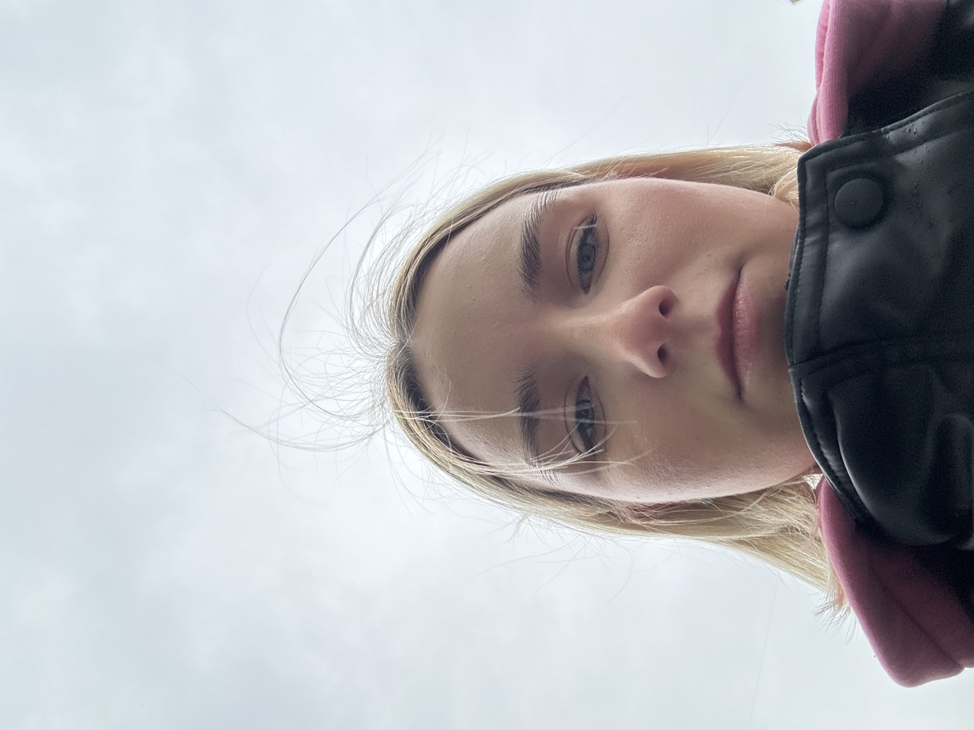 A close-up, low-angle shot reveals a young woman with light blonde hair, wearing a black leather jacket over a pink hooded sweatshirt. She is facing downwards toward the camera, her gaze directed toward the viewer. The background consists of a uniformly gray, overcast sky. The location is specified as the city of Unknown, Russia, but there are no identifiable features to confirm this.