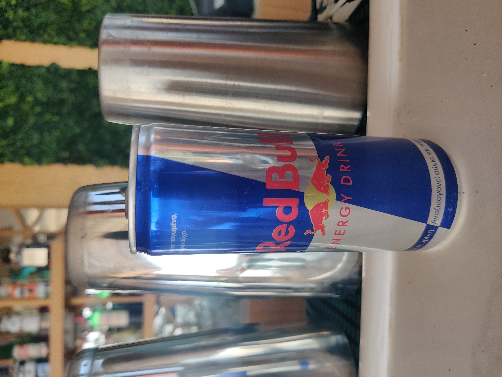 An eye-level, close-up shot focuses on a Red Bull energy drink can. The can is positioned centrally in the foreground, its blue and silver design prominently displayed. To the left and right of the can are stainless steel shakers, creating a bar-like atmosphere. In the blurred background, shelves lined with bottles of liquor are visible, suggesting a bar or restaurant setting in the city of Sithonia Municipal Unit, Greece. The lighting is bright, casting reflections on the metallic surfaces.