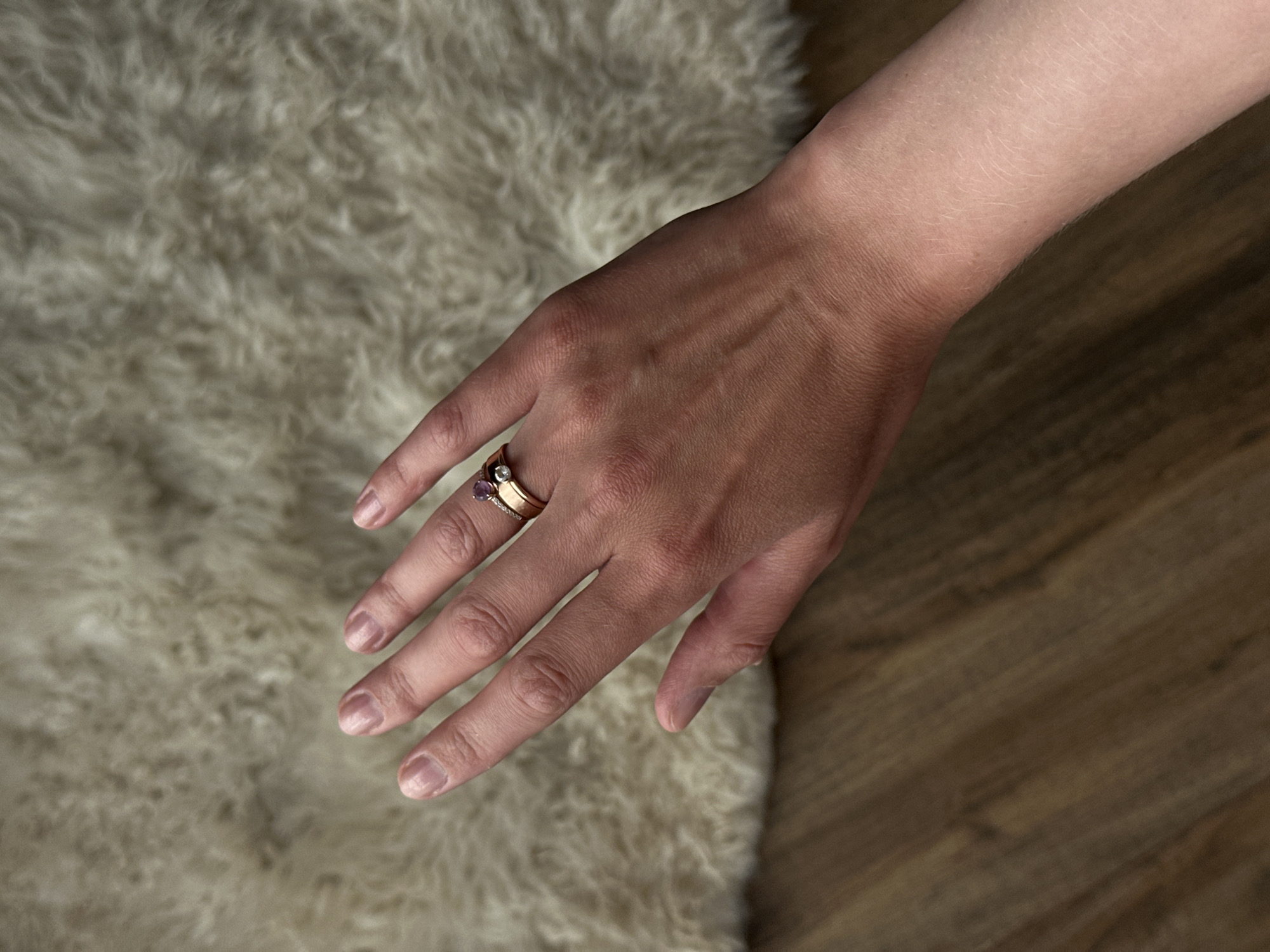 A fair-skinned hand with two gold rings (one with a purple stone and another with a white stone) on the ring finger is positioned diagonally across the image. The hand is set against a backdrop of a fluffy, off-white rug on the left and wooden flooring on the right. The location, Sepych, Russia, is not evident from the visual information provided.