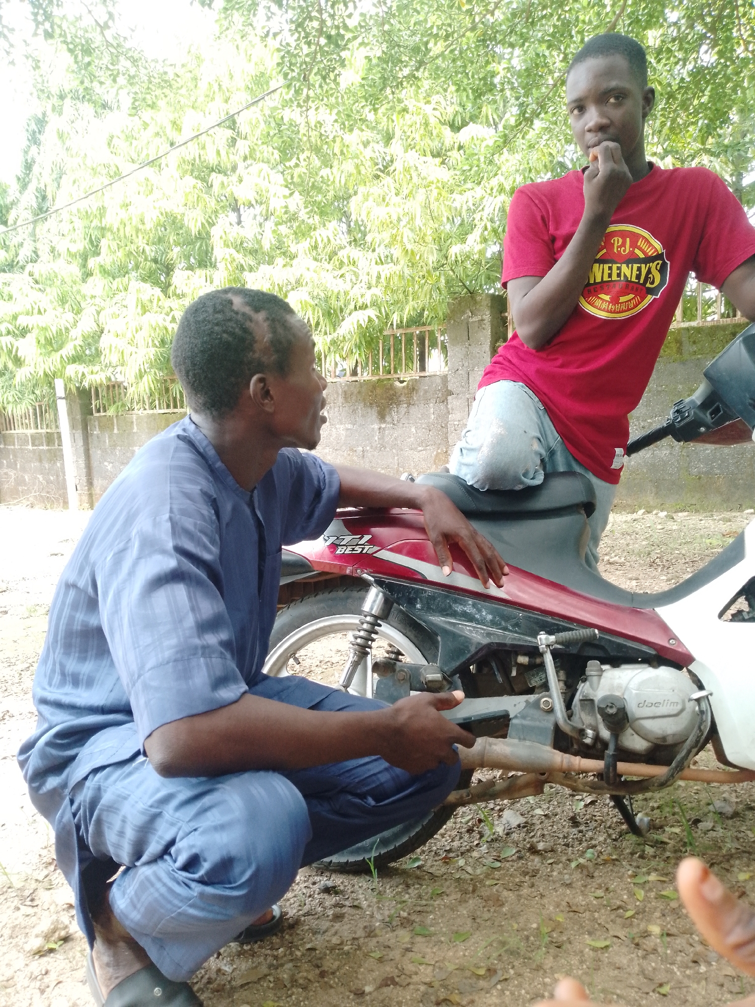 The image depicts two young men outdoors in what appears to be a semi-urban or rural setting, possibly in Akwanga, Nigeria. In the foreground, one man is squatting beside a red and white motorcycle. He is wearing a blue, loose-fitting, striped traditional garment. His left arm is resting on the motorcycle seat, and his right hand is near the engine. The second man is standing slightly behind and to the right, leaning on the motorcycle. He is wearing a red t-shirt with a yellow circular logo that reads "P.J.WEENEY'S" and light blue jeans. He is bringing his hand to his mouth as if eating or tasting something. The background shows lush green foliage from trees, a concrete wall, and a metal fence. The ground is covered in dirt, small stones, and scattered dry leaves. The lighting suggests it is daytime with overcast conditions. The overall scene is informal and casual.