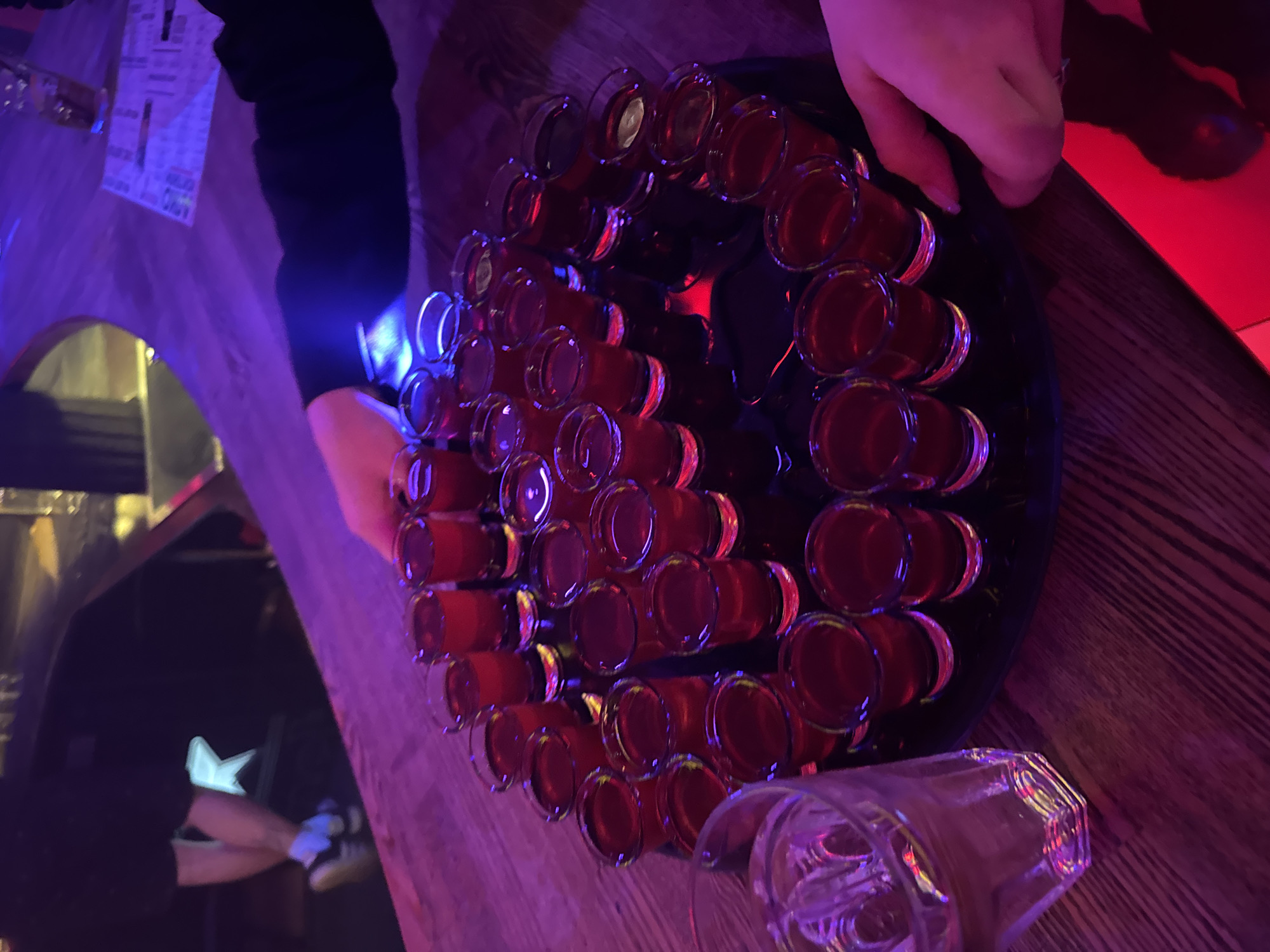 The image depicts a close-up, overhead view of a tray filled with numerous small shot glasses, each containing a dark red liquid, likely alcoholic. The tray is situated on a wooden bar counter. In the foreground, to the left, a partially visible glass of water or clear liquid is present. In the background, out of focus, a person's legs can be seen, wearing dark shorts, white socks, and sneakers. Another person's hands are visible reaching towards the shot glasses, suggesting an interaction with the drinks. The lighting is dim and colorful, with purple and red hues dominating, indicative of a bar or nightclub atmosphere. The scene is devoid of any discernible identifying features that would place it specifically within the city of Perm, Russia; it represents a generic bar setting.