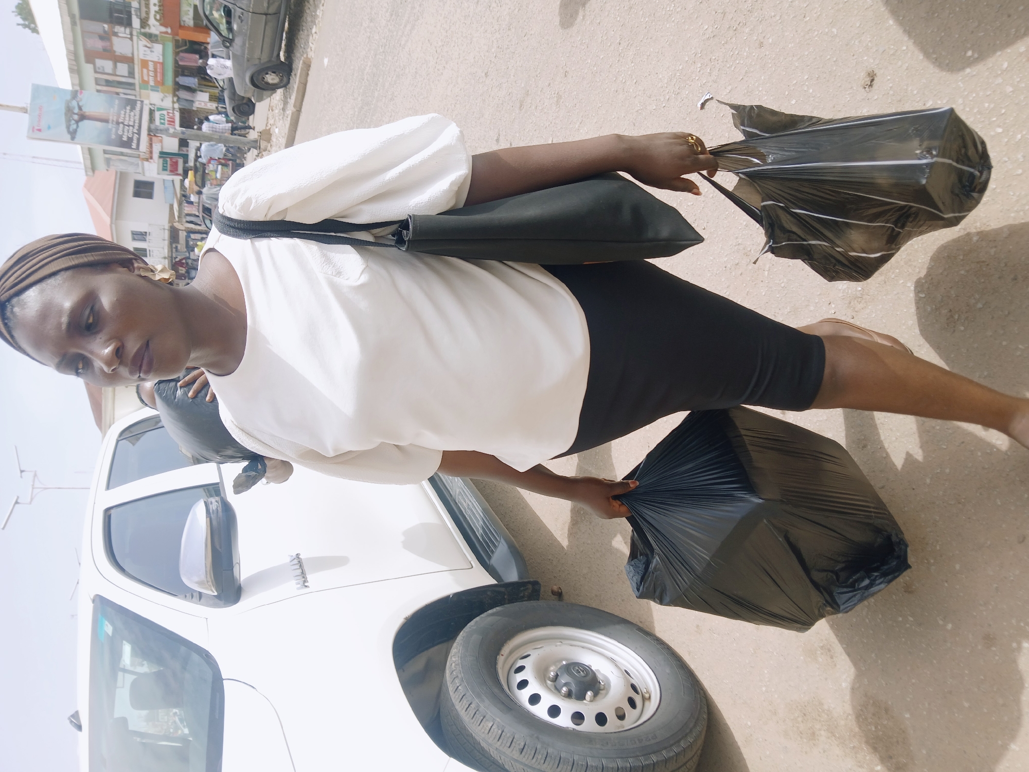 A woman stands beside a white pickup truck in Jos, Nigeria. She wears a white blouse with puffed sleeves, black shorts, and a brown head covering. She is also wearing a gold ring on her right hand. A black bag hangs from her shoulder, and she holds a black plastic bag in one hand and a black umbrella in the other. The truck, a white Toyota Hilux, is parked on a paved surface. In the background, buildings and street signs can be seen, suggesting an urban location.
