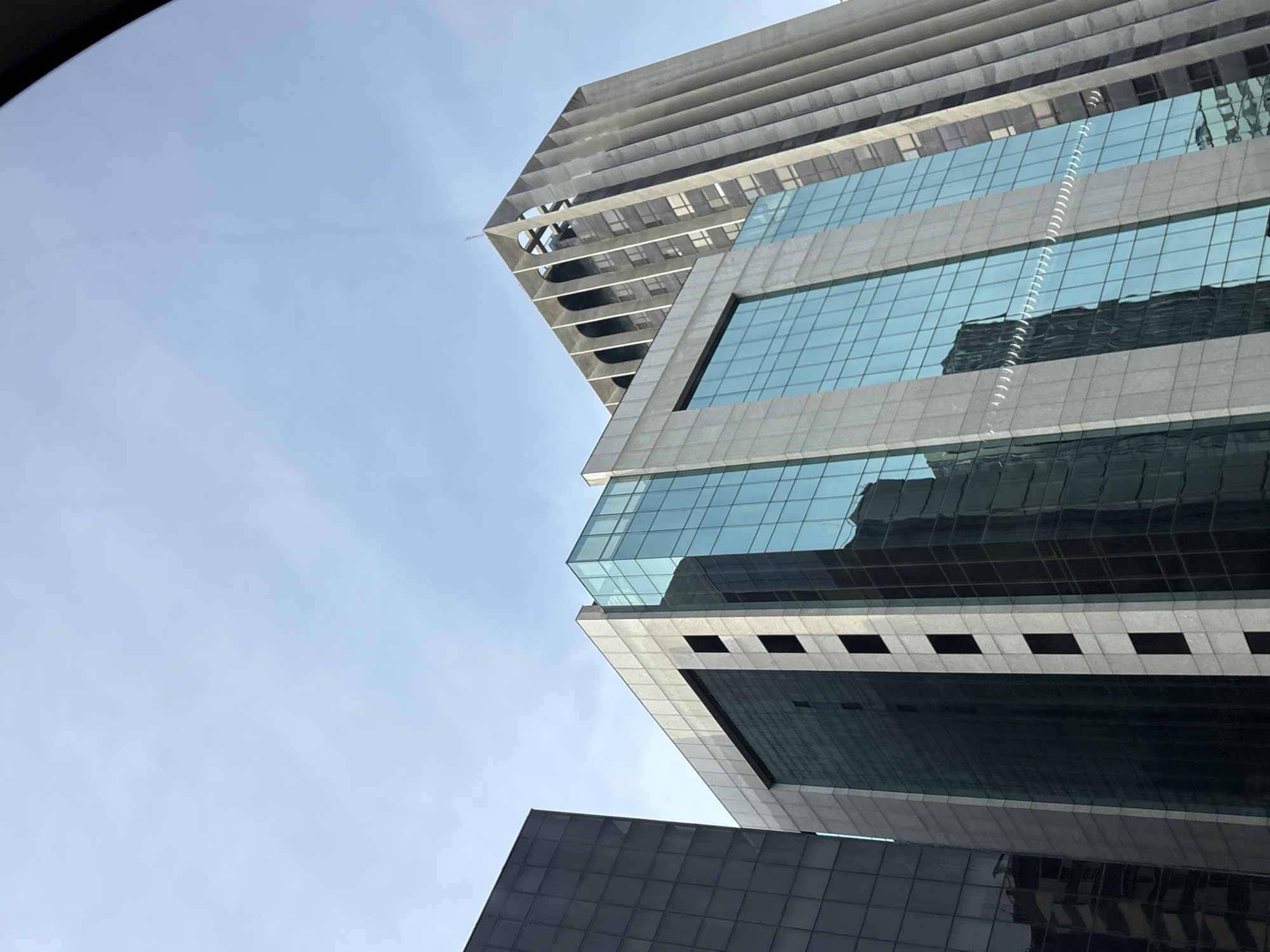 The image is a low-angle shot of modern skyscrapers in the city of São Paulo, Brazil. In the foreground, a dark-paneled building is partially visible on the left. To its right, a building with a facade of light-colored stone and turquoise glass panels dominates the center of the frame. The turquoise glass reflects the sky and the surrounding buildings. Further to the right and behind the glass-fronted building, a Brutalist-style skyscraper with a distinctive concave facade and vertical concrete elements rises. The sky above is a clear, pale blue with a few wispy clouds. The image captures a section of the urban landscape, emphasizing the architectural styles of the buildings and their relationship to the sky. There are no people or specific actions depicted in the image.