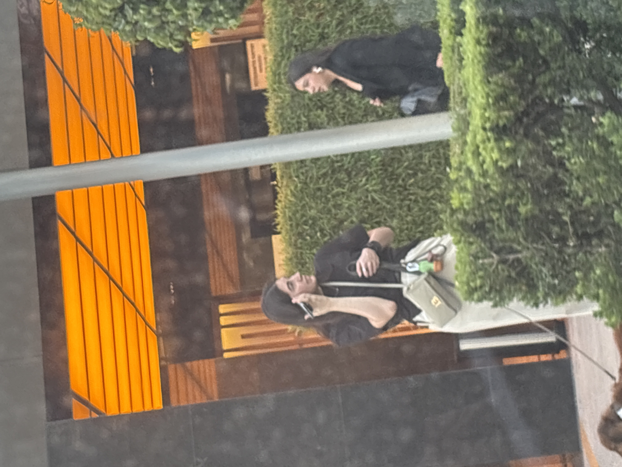 The image, if viewed upright, captures two women in an urban outdoor setting, likely in Mexico City as per the provided context. A prominent horizontal grey metal bar and a continuous line of green, well-maintained hedges divide the scene into distinct foreground and background planes.
In the foreground, a woman with dark hair, wearing a black short-sleeved top and light-colored pants or a skirt, is actively engaged in a phone call. She holds a dark smartphone to her right ear, looking slightly upward and to her left. A light green shoulder bag, with the letters "ET" faintly visible on its side, hangs across her body. A dark watch or bracelet is visible on her left wrist.
In the background, behind the hedges, another woman with dark hair is partially visible. She is dressed in a black top or dress and appears to be looking downwards or to the side. A white earpiece, resembling an AirPod, is noticeable in her left ear.
The backdrop for both women consists of dark building facades interspersed with striking, bright orange slatted panels or blinds, which create strong vertical lines and warm reflections. Additional architectural elements, possibly made of wood, are also visible. The lighting in the scene is soft and ambient, suggesting daytime, possibly late afternoon. The overall impression is a casual, urban moment.