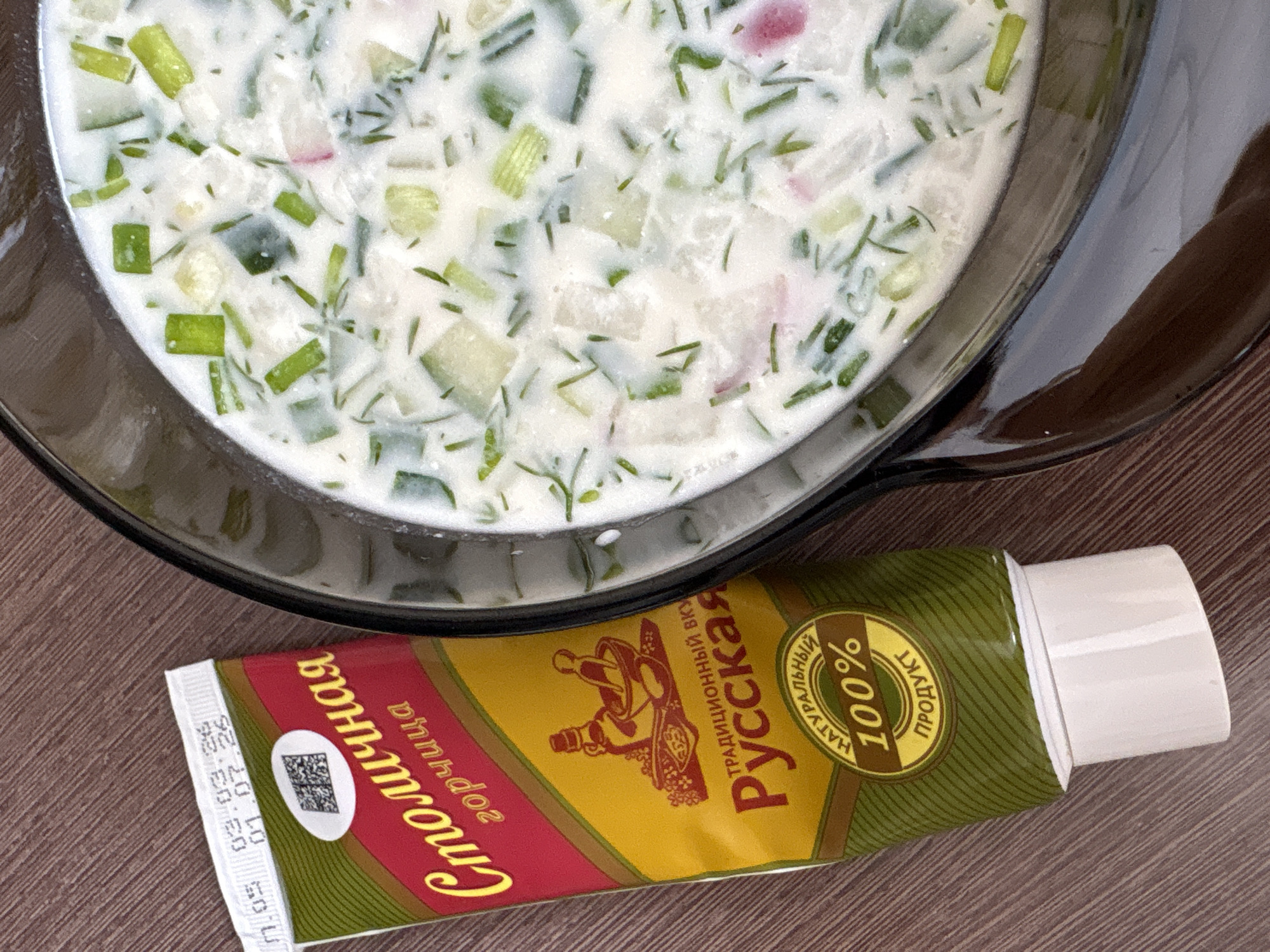 A top-down shot reveals a bowl of okroshka, a cold soup, next to a tube of Russian mustard. The soup contains ingredients like diced cucumbers, green onions, dill, and possibly radishes, suspended in a white, milky broth. The bowl is black with a slight sheen. The tube of mustard is green, yellow, and red, with Russian text and a label indicating it is a 100% natural product. A QR code is visible on the mustard tube. The background is a wooden surface. There's no indication of Ust-Ilimsk, Russia.