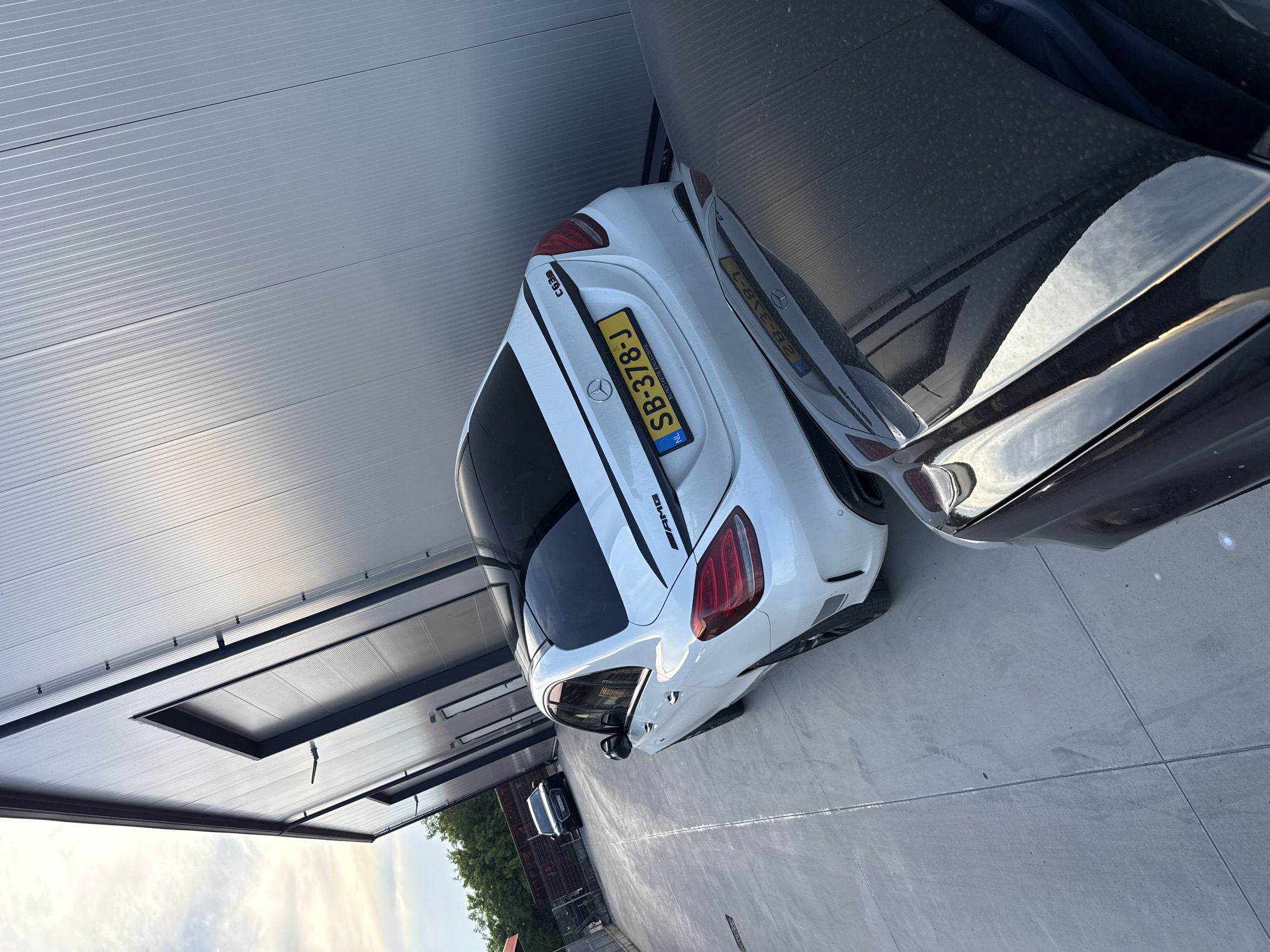 The image shows the rear of a white Mercedes-Benz C63 AMG sedan parked on a concrete surface in front of a modern industrial building with a corrugated metal exterior. A black car is partially visible in the foreground. In the background, another black car is parked further down the lot. The sky is visible with some clouds, and there are trees to the left. The license plate on the white Mercedes reads "SB-378-J". The building appears to be a commercial or industrial facility, possibly a warehouse or workshop. The overall scene suggests an outdoor parking area associated with a business in an urban or suburban setting.