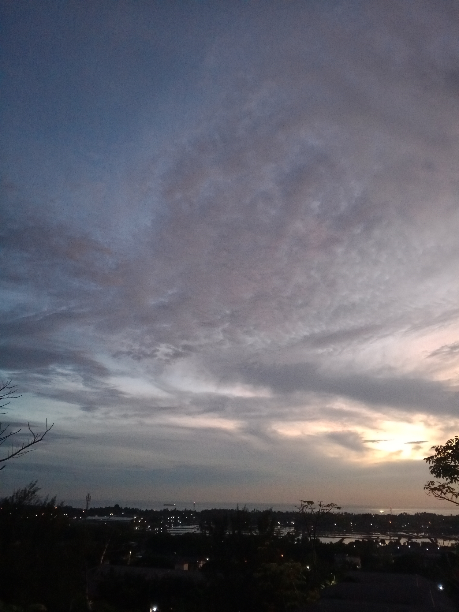 The image depicts a twilight or dusk scene in Lhokseumawe, Indonesia. The sky is dominated by a mixture of blue and gray clouds, with a slight gradient of orange and pink hues near the horizon suggesting the sun has recently set or is about to rise. The cloud formations are varied, with some appearing wispy and others more textured.
Below the sky, a dark silhouette of trees and vegetation forms the foreground. Scattered points of light indicate the presence of city lights in the distance, likely from buildings and infrastructure within Lhokseumawe. A body of water, possibly the sea, is visible between the treeline and the horizon, reflecting some of the remaining light from the sky. There are no visible people or explicit interactions in the scene.