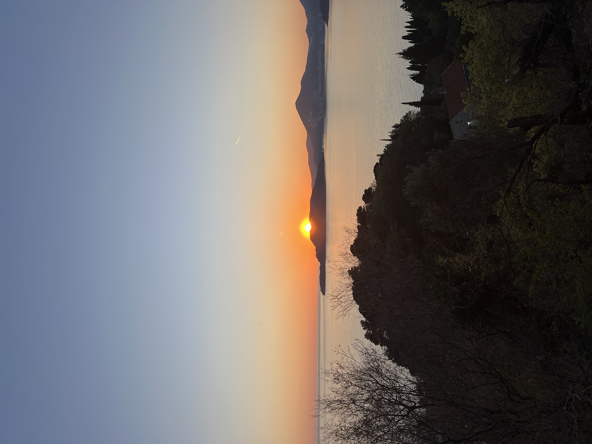 The image depicts a sunset scene in Sveti Stefan, Montenegro. The sun is setting behind mountains over water. The sky transitions from blue to orange near the horizon. The right side of the image is dominated by foliage, including trees. A jet contrail is visible in the sky. A building with a red roof is visible in the foliage on the right.