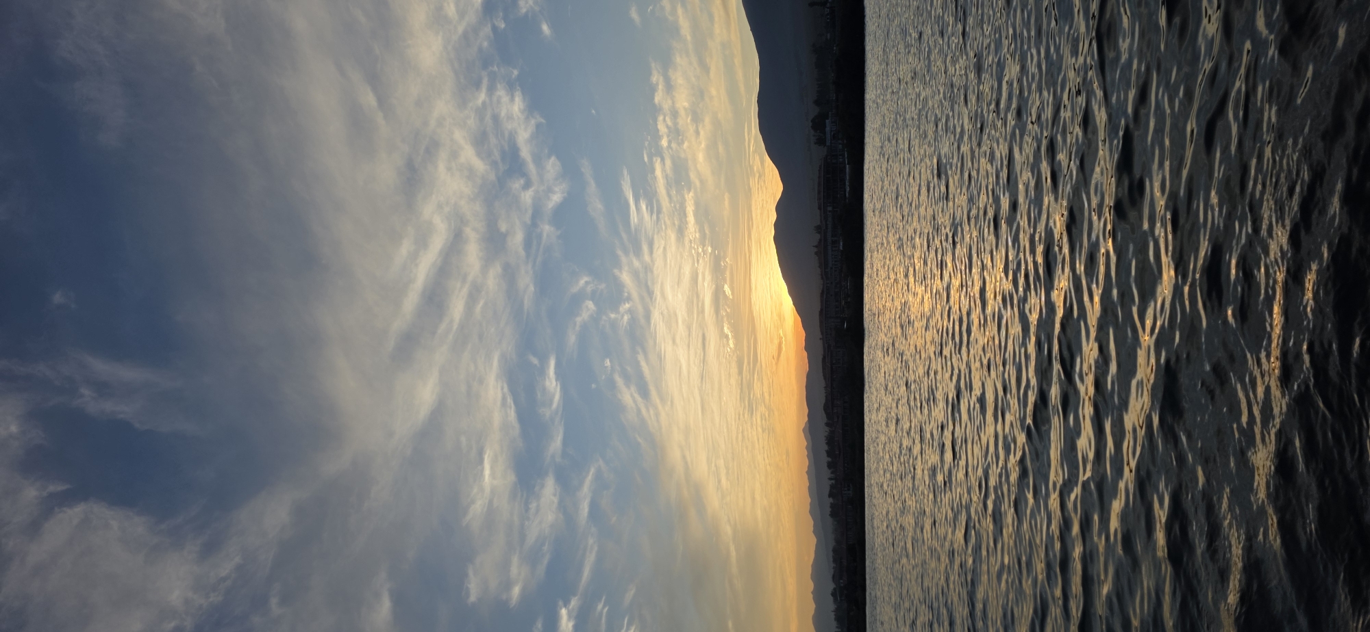 This is a vertical, outdoor, full shot of a body of water at sunset. The sun is setting behind a line of mountains, casting a warm orange glow across the sky and reflecting on the water's surface. The water itself is rippled, with darker tones of blue and grey dominating the foreground. In the distance, silhouetted against the sunset, is a town or city with buildings and trees. The sky above is a gradient of blue, transitioning from a deep blue at the top to a lighter orange and yellow near the horizon, with wispy white clouds scattered across it. The image appears to capture a serene moment at dusk.