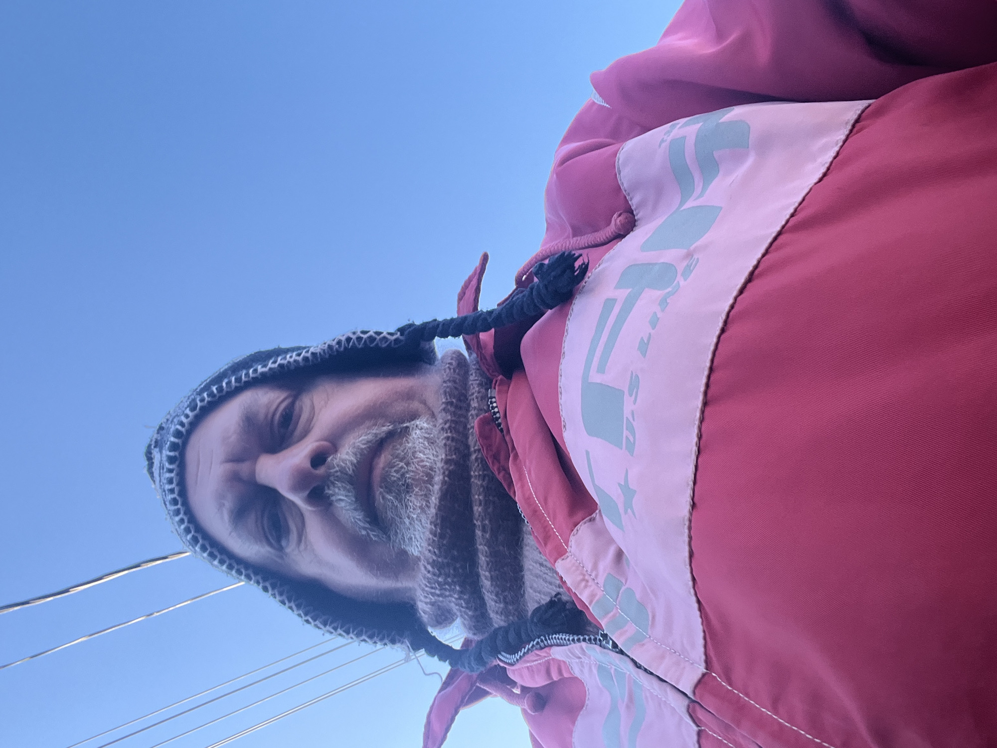 A low-angle shot depicts a middle-aged man with a white beard wearing a knit cap and scarf, as well as a pink and red jacket. The background is a clear blue sky. Power lines are visible. The location is not specified, but the text requests the location to be Podolsk, Russia, despite it not being apparent from the image.