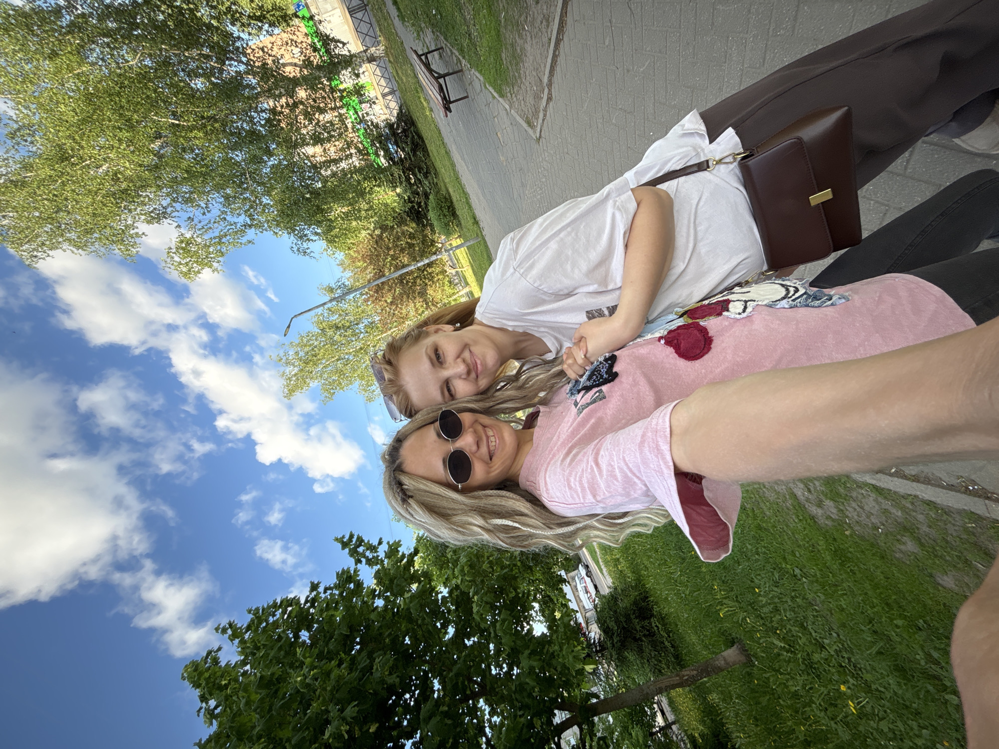 Two women are taking a selfie outside in Rusanovo, Russia. The woman closer to the camera has blonde hair and is wearing sunglasses and a pink shirt. The woman behind her has reddish-blonde hair and is wearing a white shirt. They are holding hands and smiling. A brown purse is on one of their laps. Behind them are trees, grass, a sidewalk, and a cloudy blue sky.