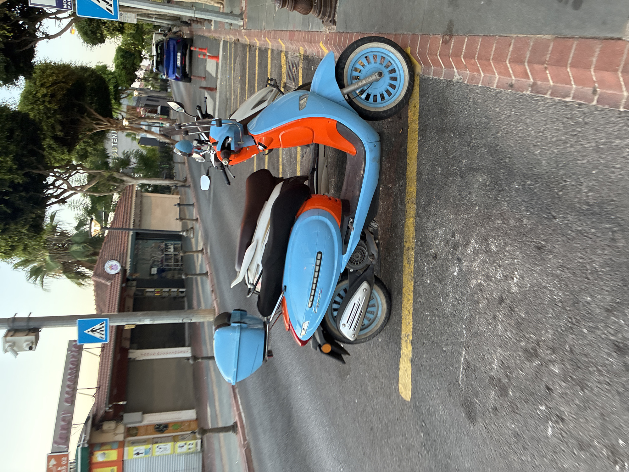 The image depicts a street scene in Alanya, Turkey, likely during the late afternoon or early evening as indicated by the warm lighting. The primary subject is a bright blue and orange scooter parked on the side of a paved road. The scooter is positioned in front of a row of shops, some of which have their shutters down, suggesting it might be after business hours.
In the foreground, the scooter is detailed: it has a blue top case, a dark seat with what appears to be another seat behind it, and a distinctive blue and orange body. The front wheel is blue with white wall tires. The road has yellow markings, including a continuous line along the edge of the sidewalk and a dashed line further into the road.
The background shows a street lined with trees and buildings. A blue car is parked further down the street, and several orange traffic cones are visible. Pedestrian crossing signs are also present on utility poles. The architecture of the buildings appears consistent with a Mediterranean or Turkish coastal town. The overall environment is urban, with a mix of commercial establishments and street furniture. No people are directly visible in the image, but the presence of parked vehicles and shops implies a populated area.