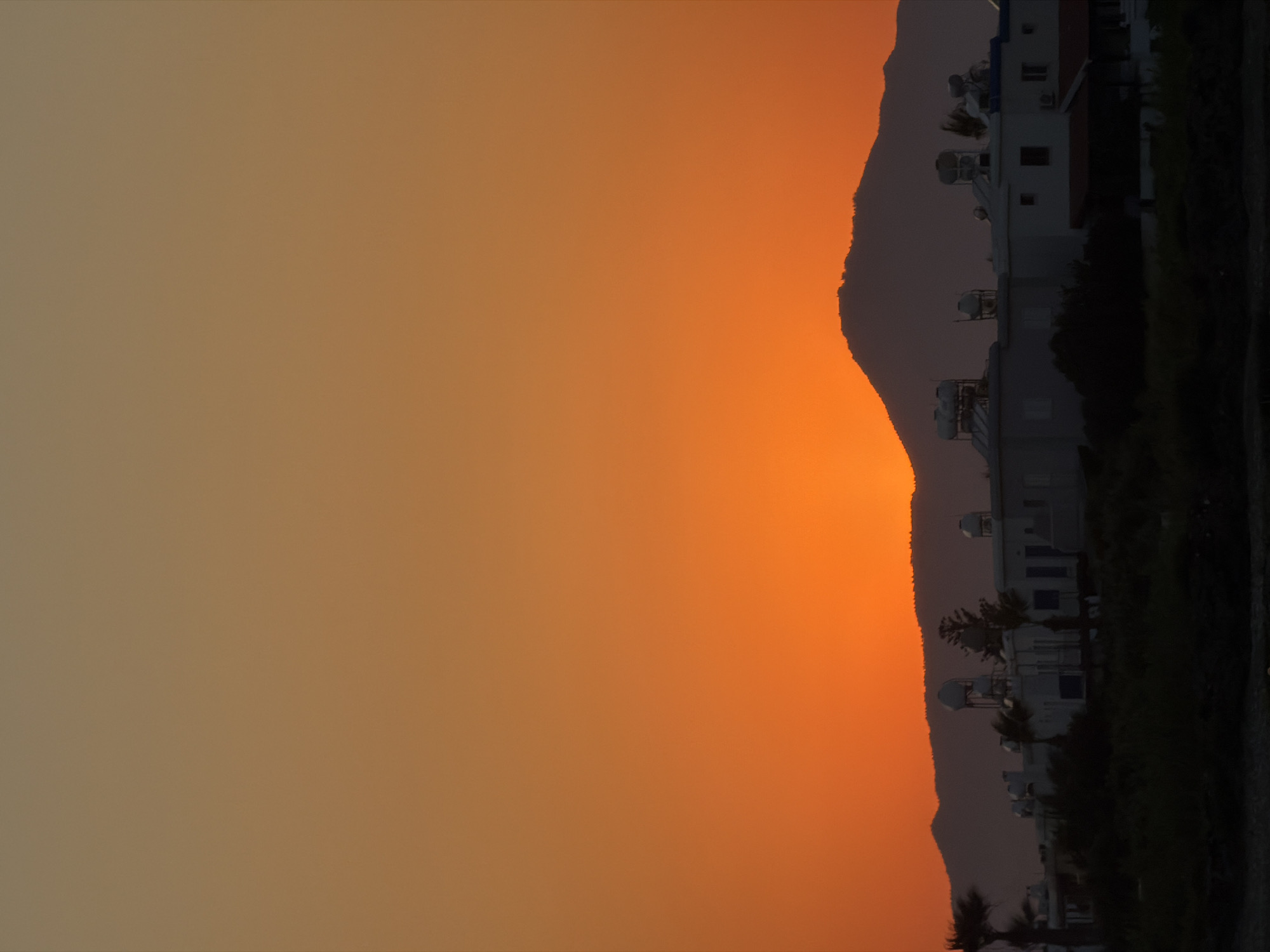 This image depicts a sunset scene in Meneou, Cyprus, as indicated by the landscape and architecture. The dominant feature is the vibrant orange and yellow gradient of the sky, characteristic of a sunset. Silhouetted against this sky are undulating hills or mountains, forming a dark, jagged line across the mid-right portion of the frame.
In the foreground, to the right of the sunset, is a row of buildings. These appear to be modern structures, possibly apartments or small hotels, with white facades, blue-framed windows, and red-tiled roofs visible on some. Several solar water heaters are mounted on the rooftops, a common sight in Mediterranean regions. Trees and foliage are scattered around and in front of the buildings, adding a touch of greenery against the dark ground. The overall impression is one of a tranquil evening in a coastal or semi-coastal area. The light suggests it is either sunrise or sunset, with the warm colors strongly pointing towards sunset. The overall mood is peaceful and serene. There is no visible text in the image.