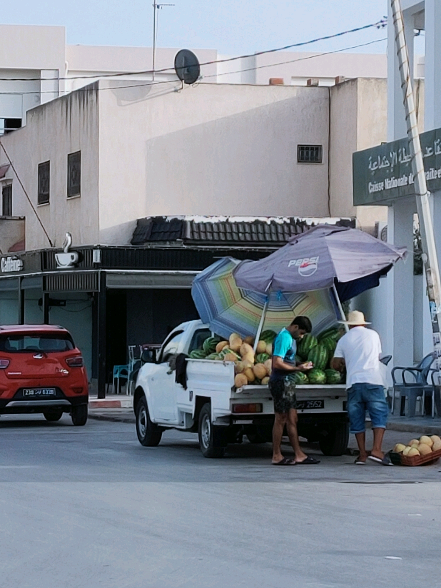 This image depicts an outdoor scene in an urban environment, likely in Unknown, Tunisia, where two men are interacting near a white pickup truck laden with watermelons and other produce. The truck is parked on the side of a street, with a red vehicle visible in the foreground to the left.
The pickup truck's bed is filled with a large quantity of watermelons, some of which are being attended to by the two men. One man, wearing a bright blue shirt and dark patterned shorts, is bent over, possibly examining or arranging the fruit. The other man, dressed in a white t-shirt and blue denim shorts, is standing with his back to the camera, wearing a straw hat. He appears to be facing the truck, and his interaction with the other man is unclear.
A large, colorful umbrella, branded with "Pepsi," is propped over the truck's cargo, providing shade. In front of the truck, to the right, a basket filled with what appear to be melons or similar fruits is placed on the ground.
In the background, buildings are visible, including a structure with a sign that reads "Caisse Nationale de S. de T." and Arabic script above it. A cafe with a sign that says "Caffeine" is also present. The overall setting suggests a street-side market or vendor stall. The lighting indicates it is daytime, possibly late afternoon given the shadows. The ground is paved, and the overall atmosphere appears to be a typical street scene in a city.