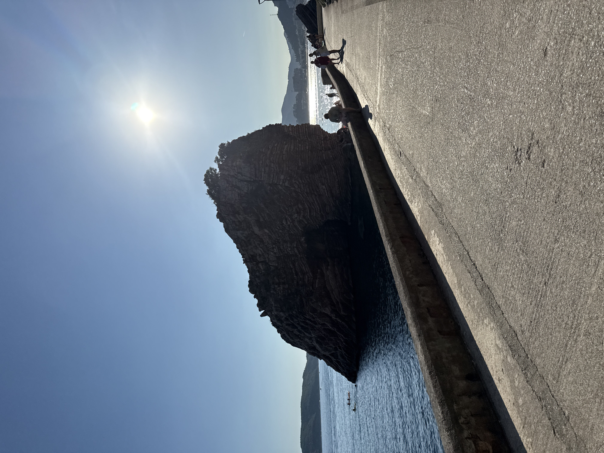 A rocky island with some vegetation on top sits in the clear blue water of the Adriatic Sea, near the coast of Rafailovići, Montenegro. The sun, bright and high in the sky, casts a glare that obscures details in the upper right portion of the image. In the foreground, a paved promenade runs along the water's edge, bordered by a low, weathered concrete wall. Several people are visible on the promenade; some are walking, while others are sitting on the wall. A few individuals are in the water, appearing to swim or float. The background shows a hilly coastline with buildings lining the shore, suggesting an urban or semi-urban setting. The water is calm, with gentle ripples reflecting the sunlight. The overall scene depicts a sunny day at the seaside.