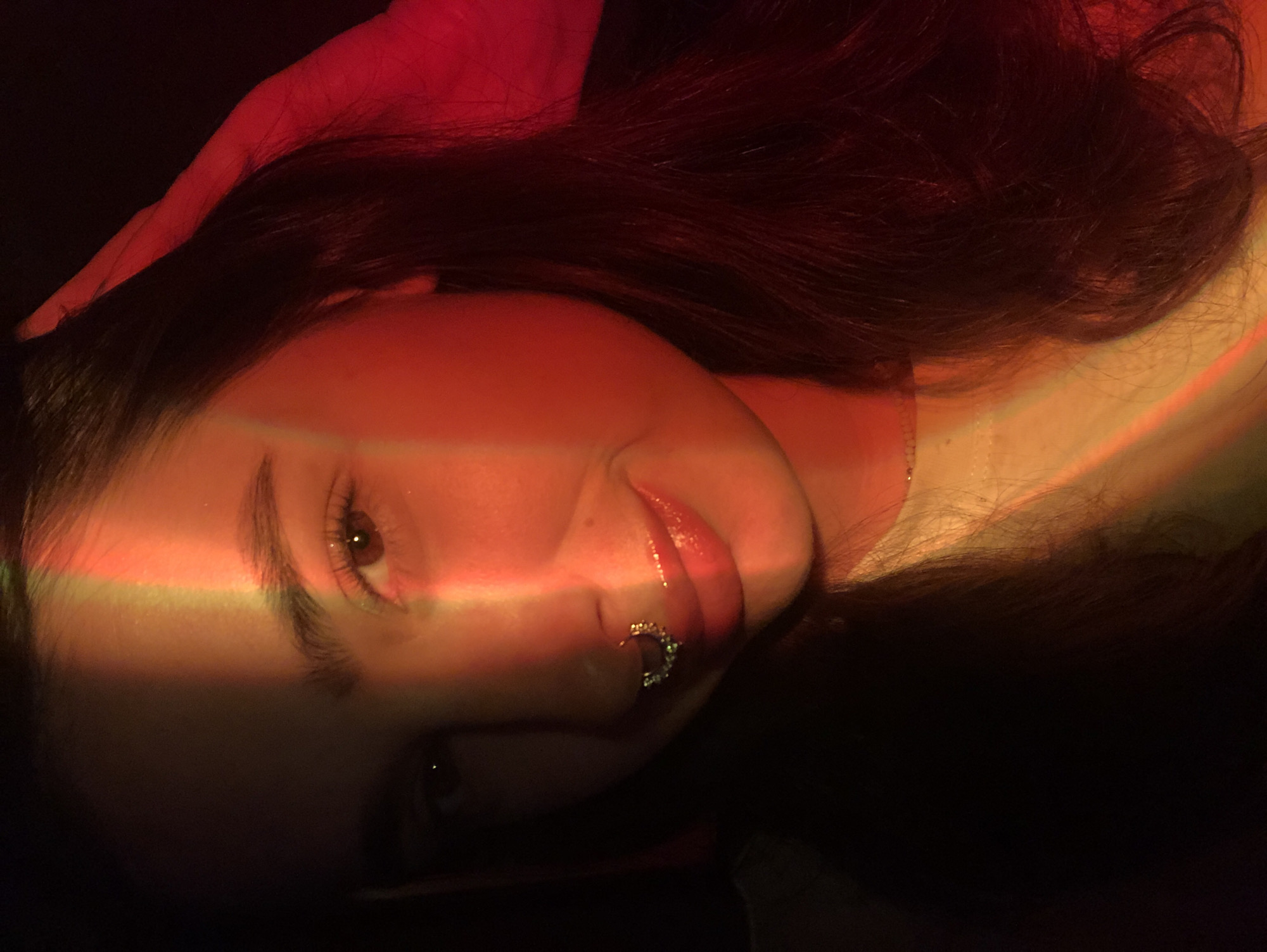 A close-up portrait of a young woman is captured, with her face partially illuminated by a rainbow-colored light source casting bands of red, yellow, and green across her features. She has long, dark hair, and is wearing a septum piercing with small, sparkling embellishments. Her right hand is raised, touching her hair near her temple. The background is dark and nondescript. The image does not provide any contextual information about the location of Noviy, Russia.